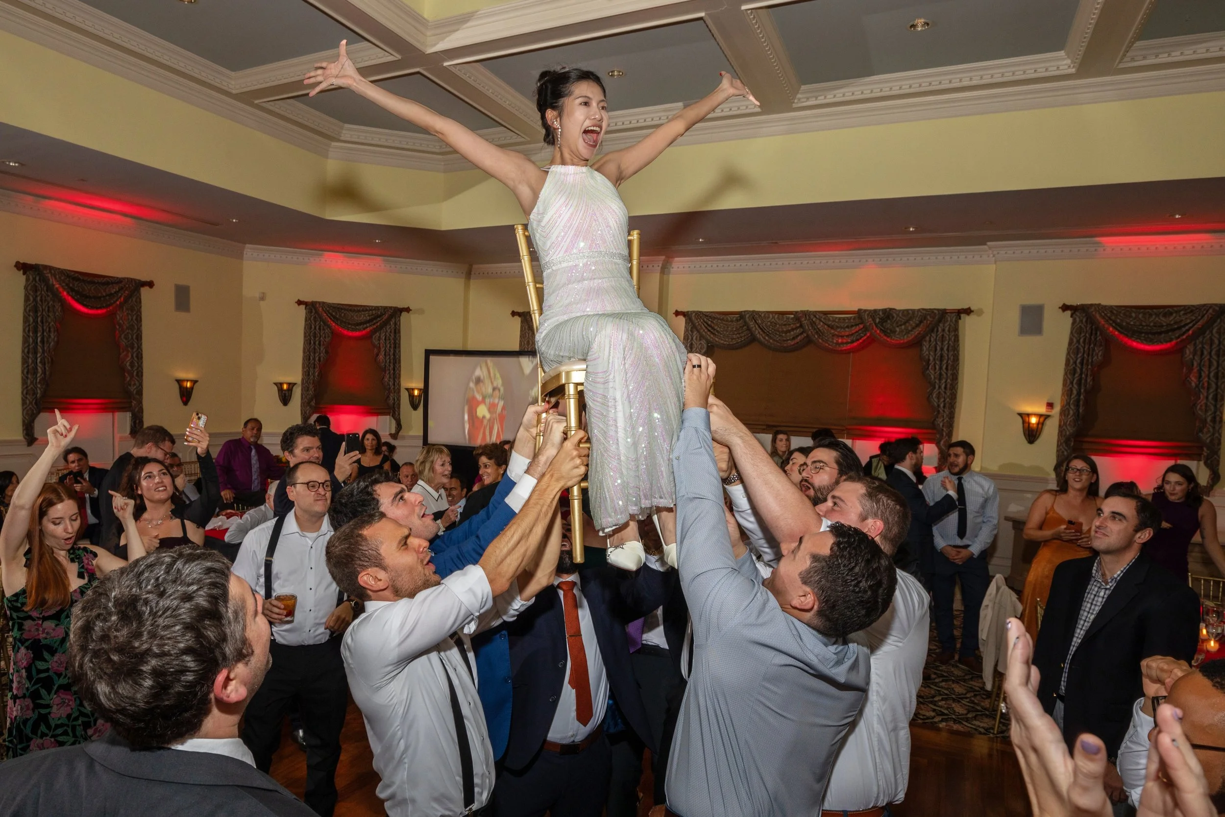 A Bat Mitzvah young woman wearing a sparkly dress sitting on a chair held up by multiple men during a celebration event, with guests dancing and taking photos in the background.