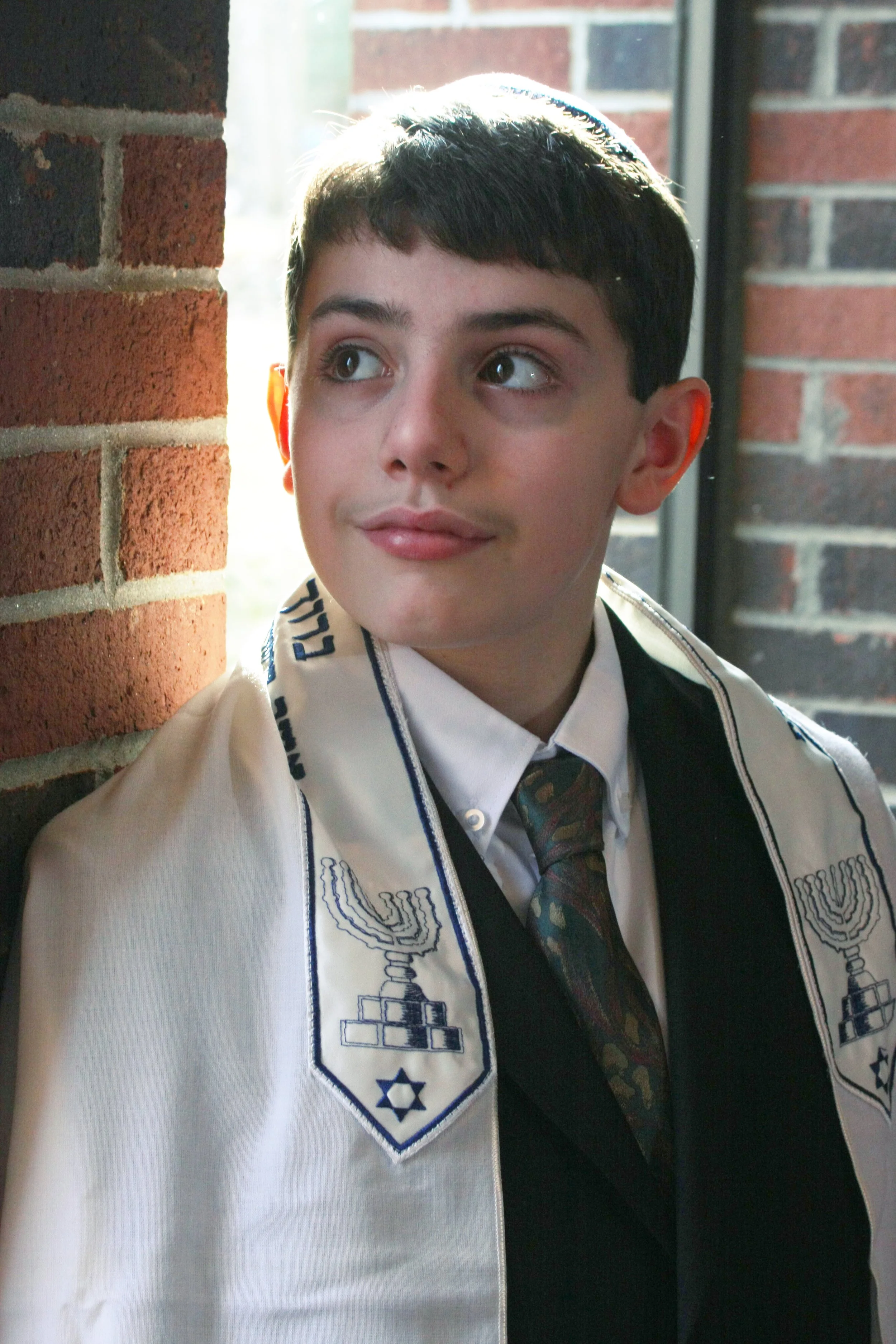 A young Bar Mitzvah boy dressed in a white shirt, patterned tie, and a tallit with embroidered menorahs and the Star of David, standing near a brick wall and a window.