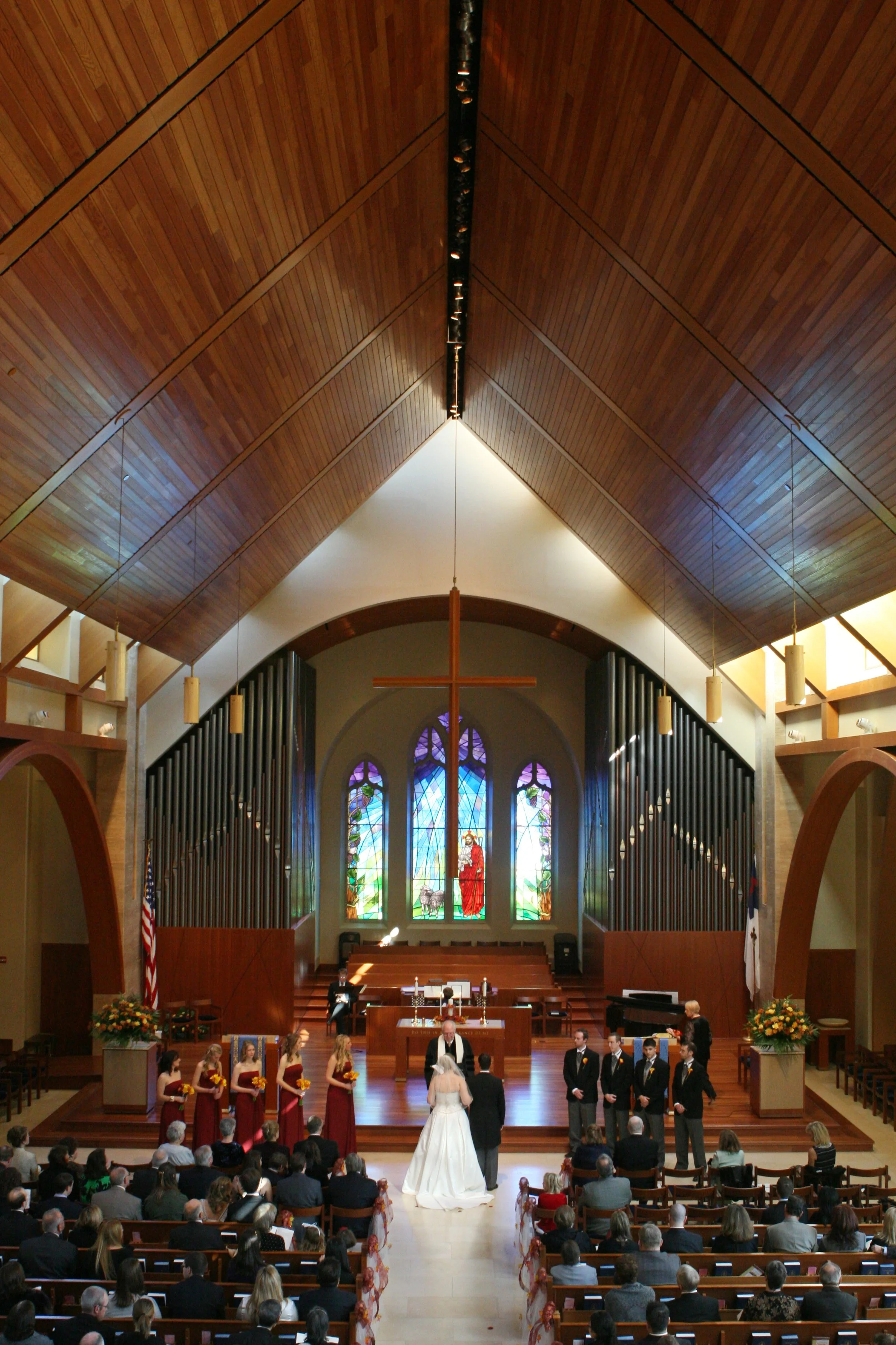 A wedding ceremony inside a church with the bride and groom standing at the altar, surrounded by bridesmaids and groomsmen, with guests seated in pews, stained glass windows, and a large wooden cross at First Presbyterian Church.
