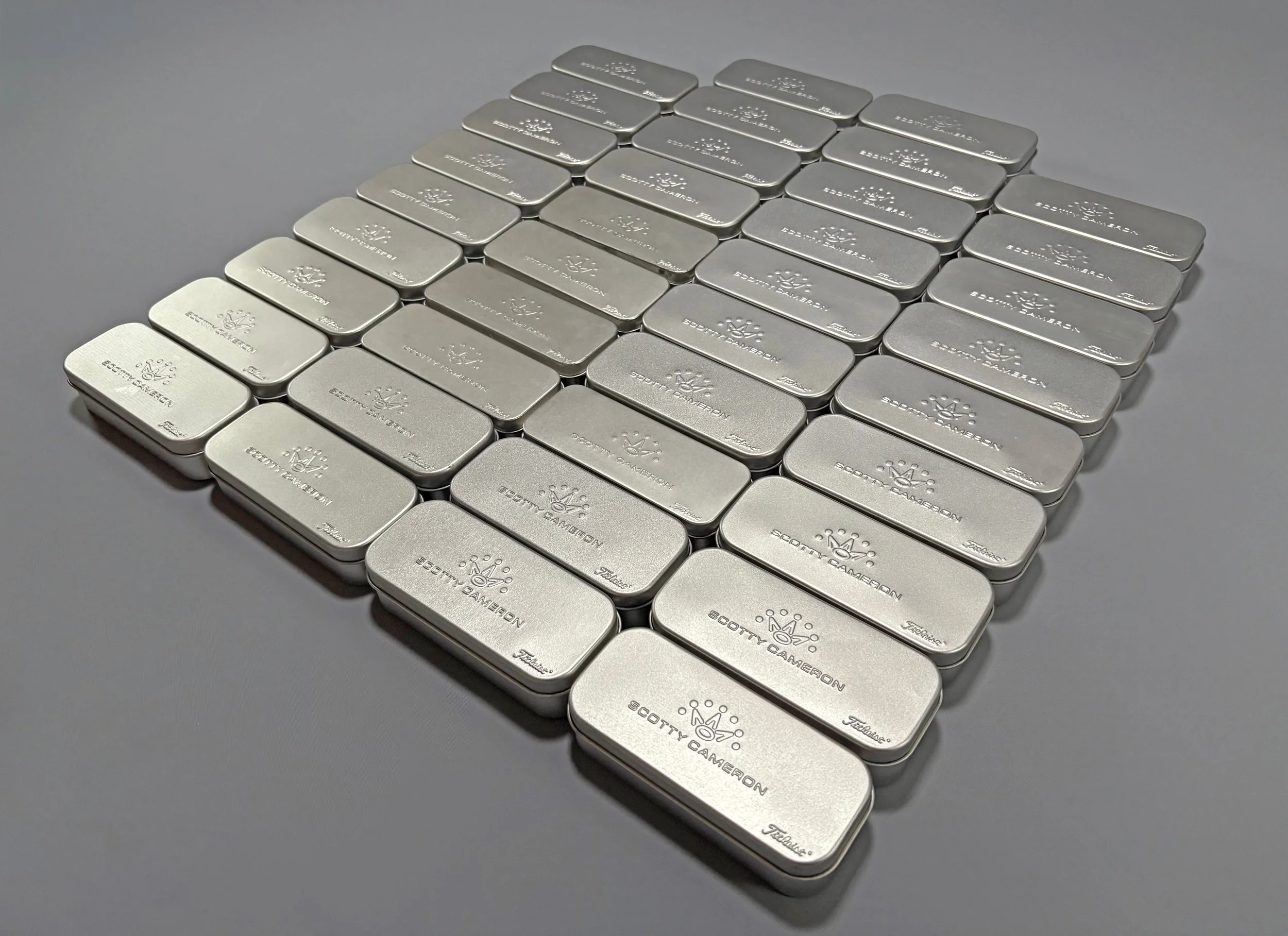 Multiple silver metal cases with engraved Scott Cameron and Titleist logos, arranged in a grid on a gray surface.