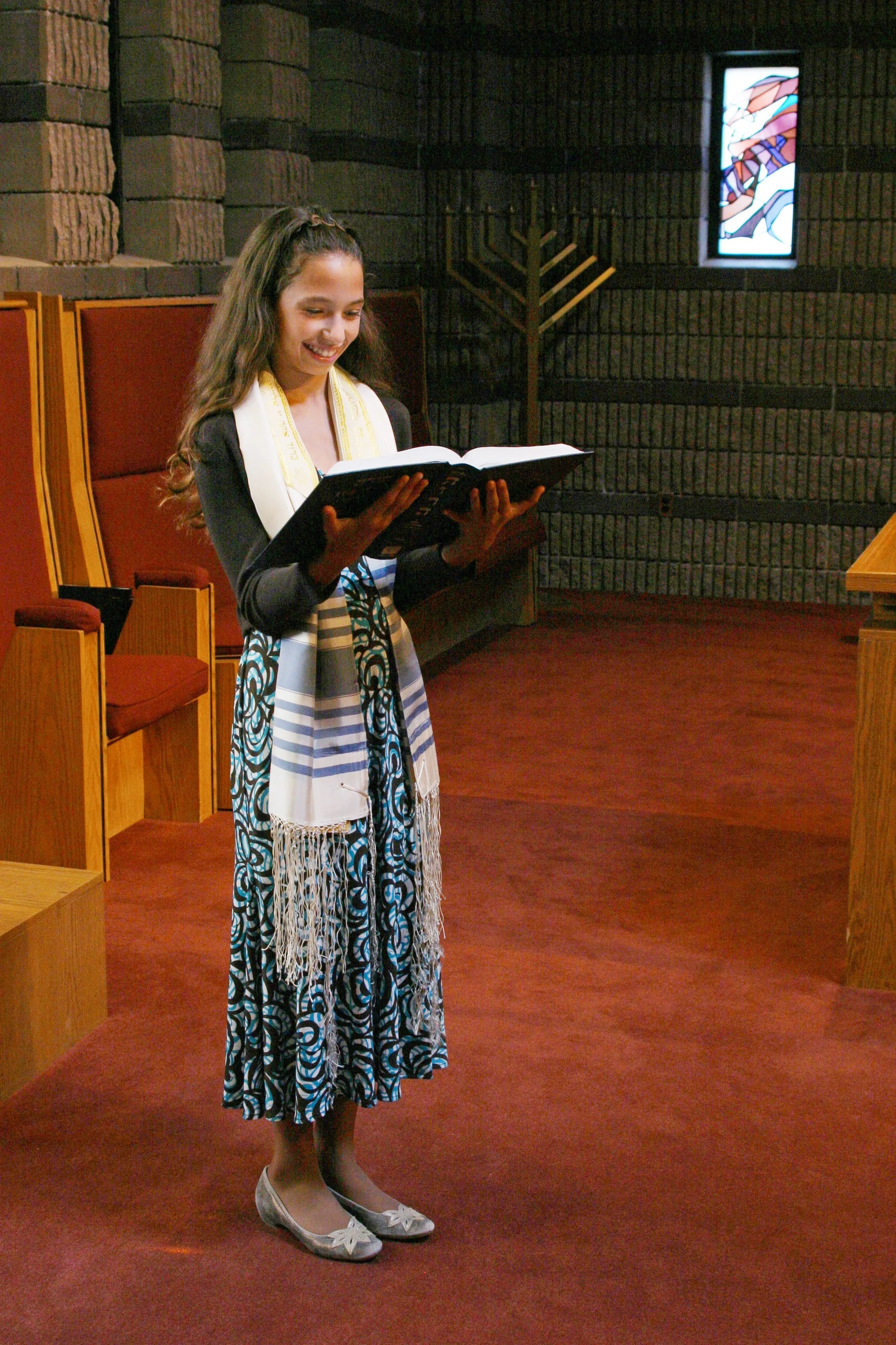 A young girl standing in a synagogue, reading from a prayer book, wearing a tallit and smiling.