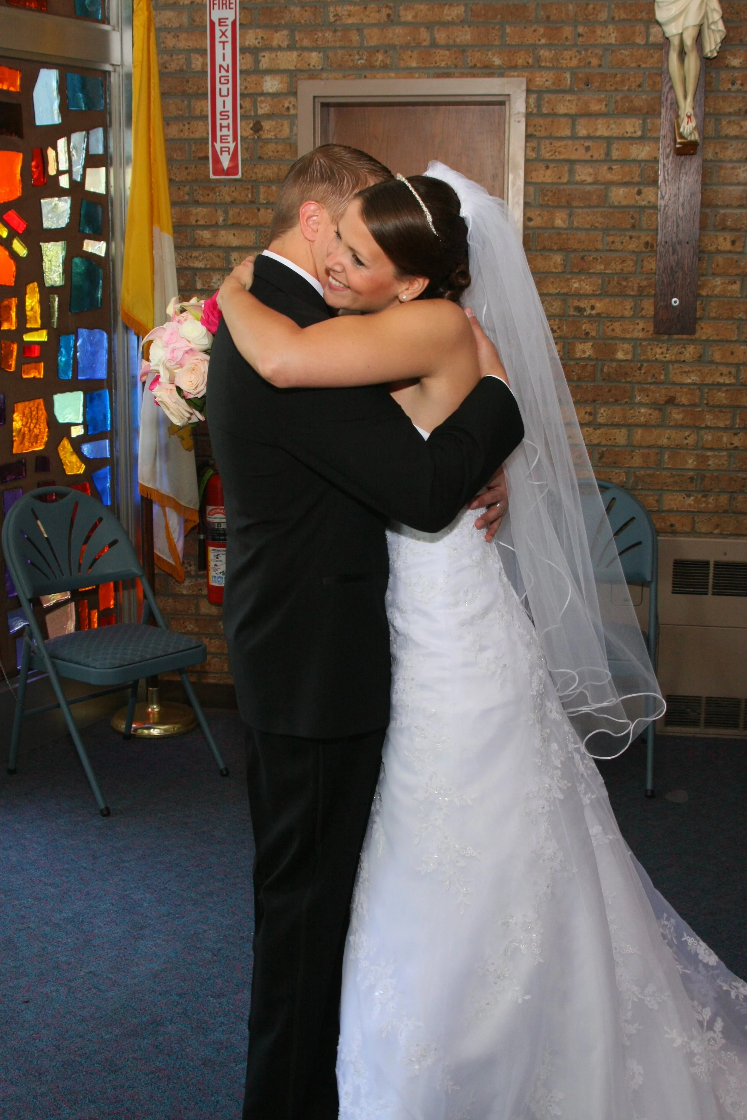 A bride and groom embracing during a wedding ceremony, with the bride in a white wedding dress and veil, and the groom in a black suit, inside a church with brick walls and stained glass windows.