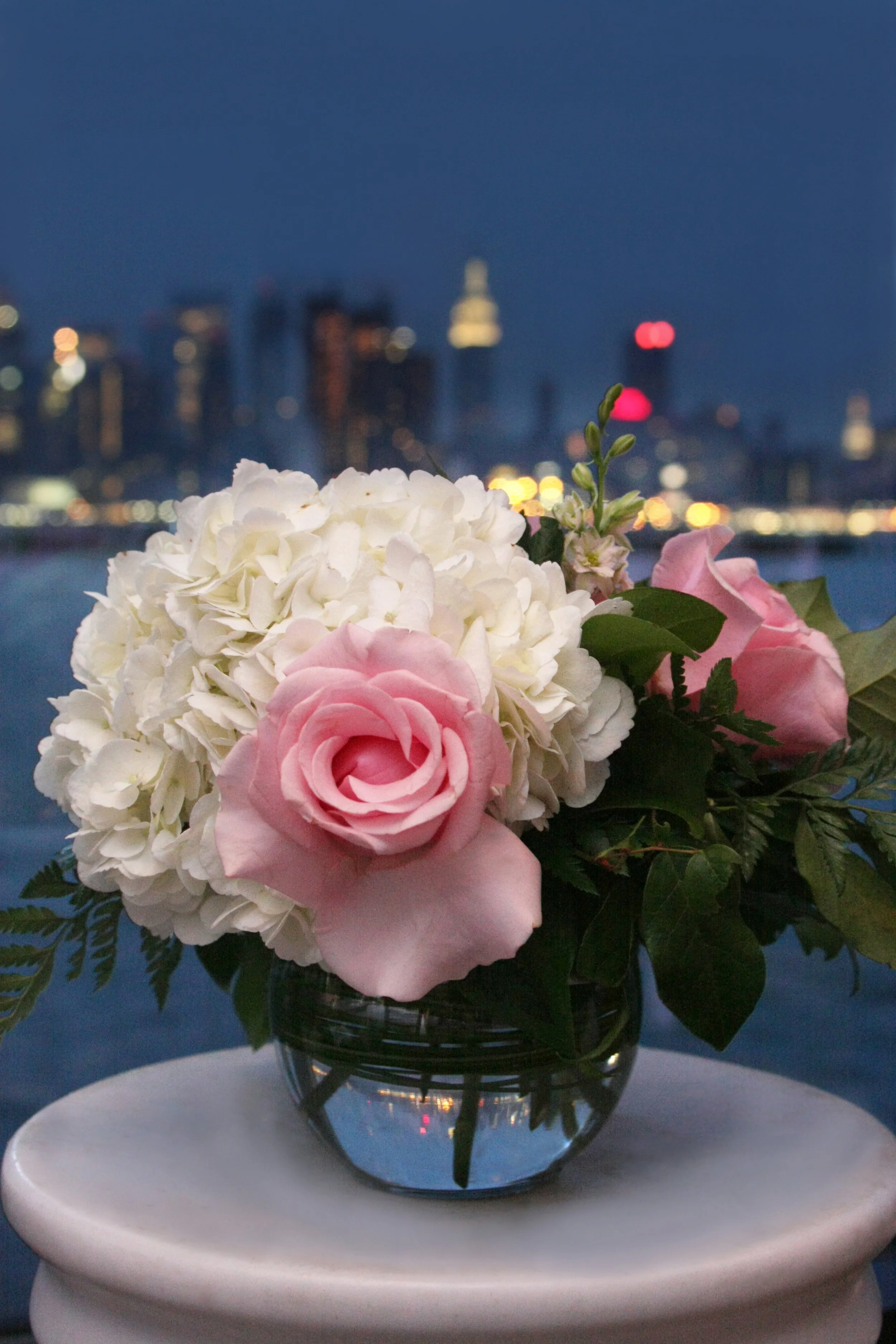 A glass bouquet of white hydrangeas and pink roses on a white surface with a city skyline and river in the background at night.