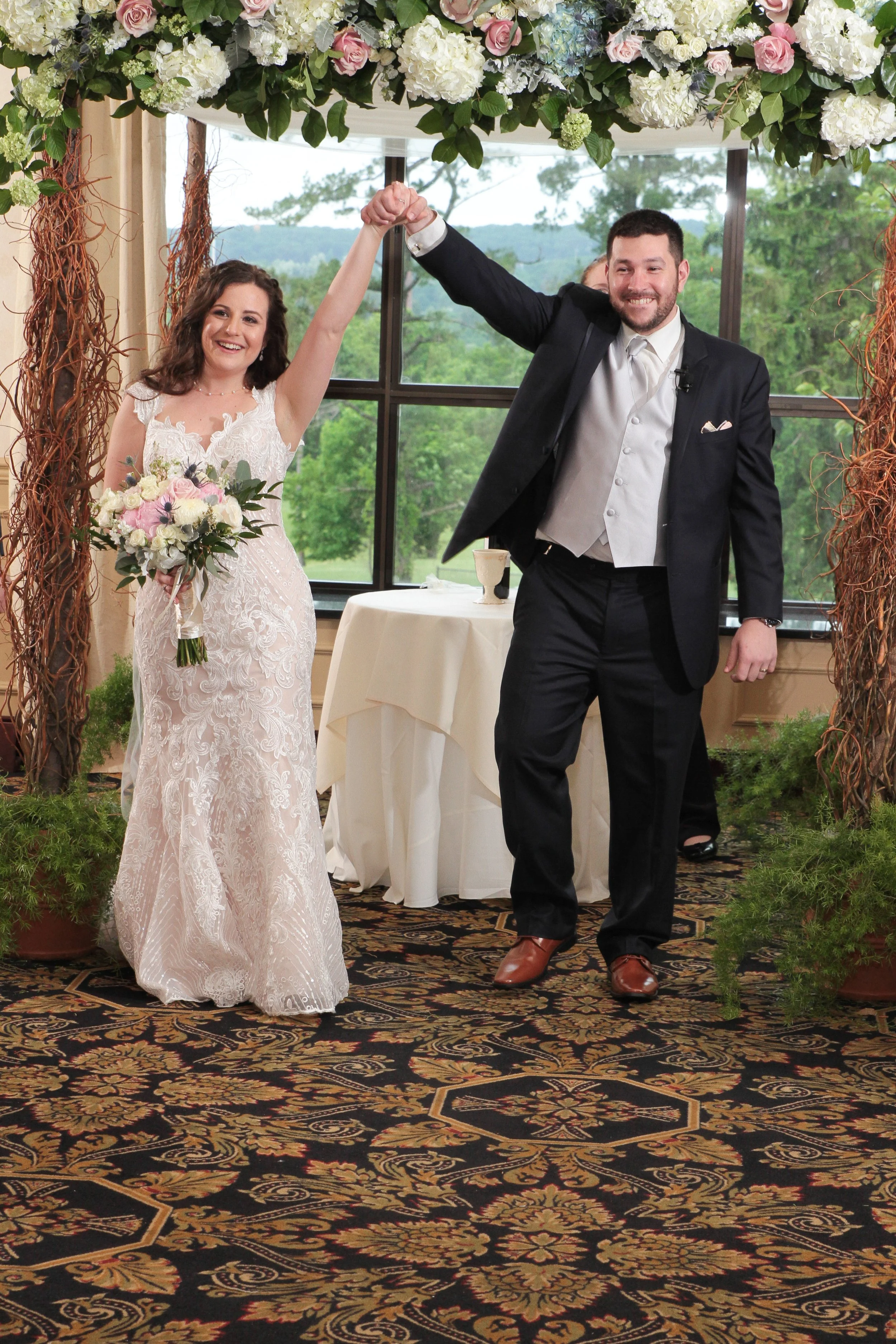 Bride and groom celebrating at their wedding reception, holding hands in the air, standing under a floral arch, with outdoor scenery visible through large windows behind them.