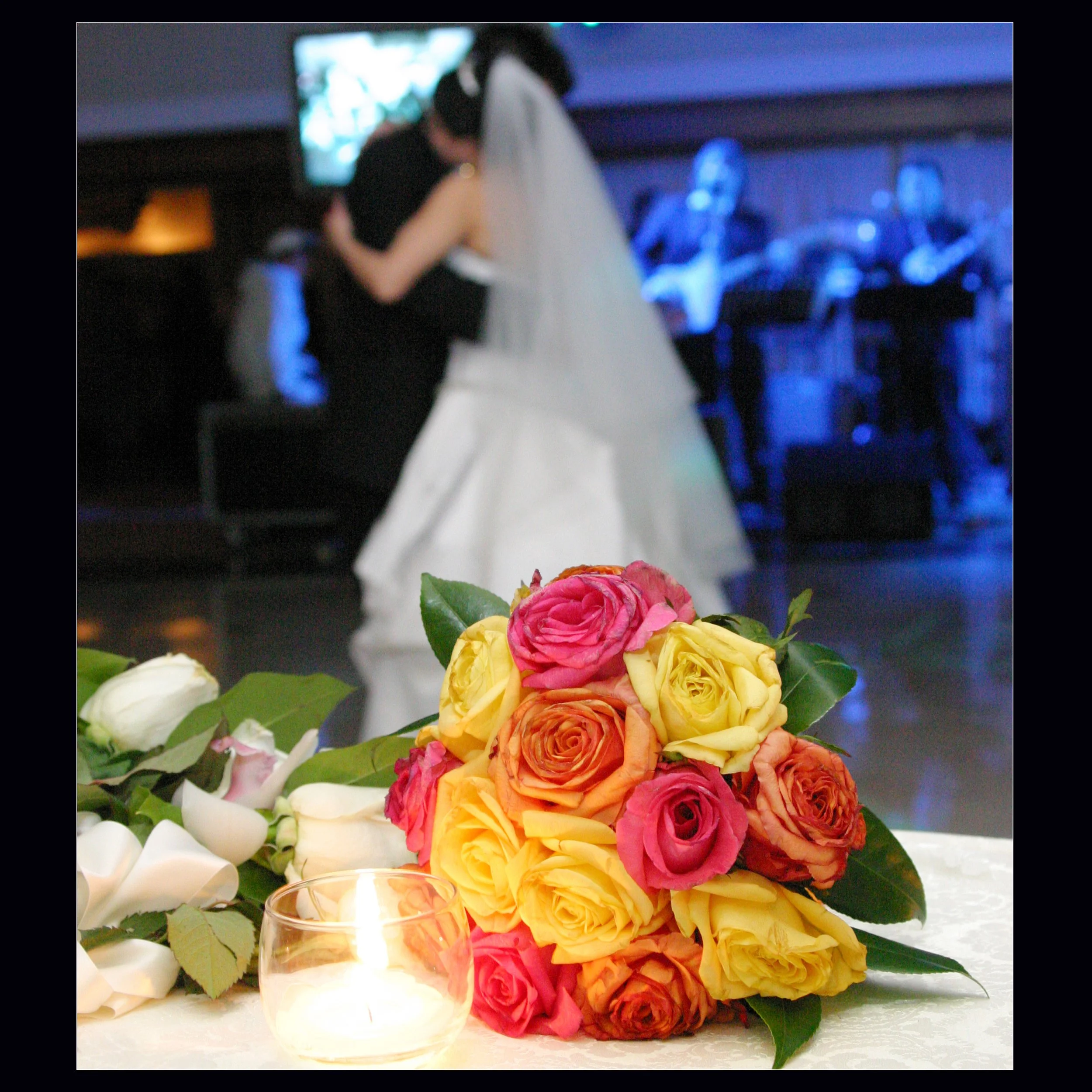 A bouquet of multicolored roses in yellow, orange, pink, and coral rests on a table, with a lit candle in a glass holder nearby. In the background, a bride and groom share a dance at their wedding reception, with a live band performing on stage.