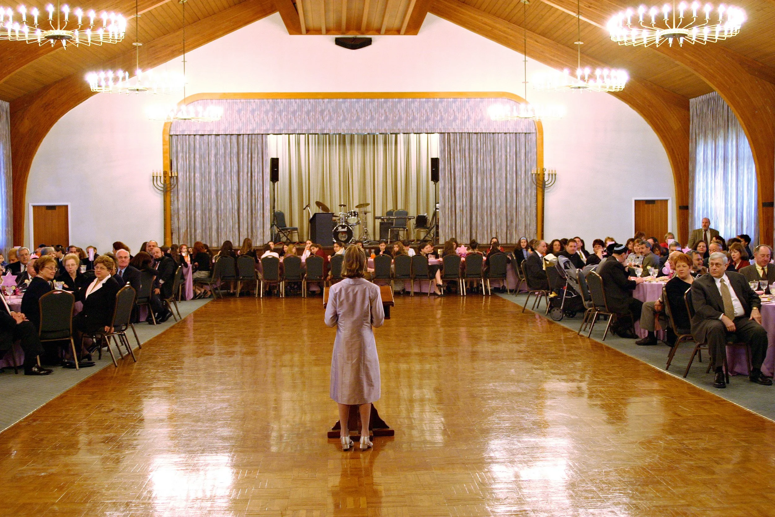 A Bat Mitzvah girl standing at a podium speaking to an audience seated at round tables in a large hall with a stage, musical instruments, and chandeliers.