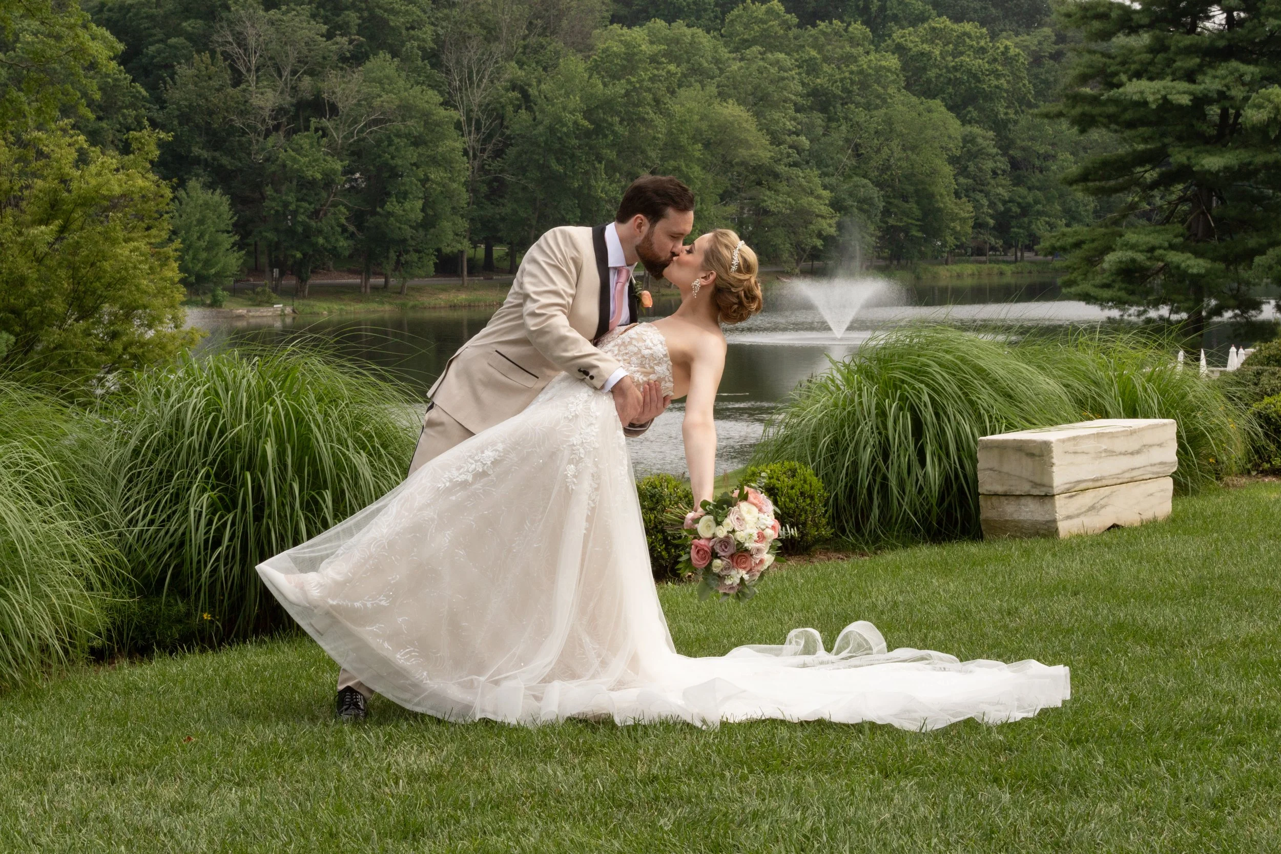 A newlywed couple sharing a kiss by a lake, with the groom dipping the bride who is holding a bouquet of pink and white roses, surrounded by lush greenery and a fountain in the background.