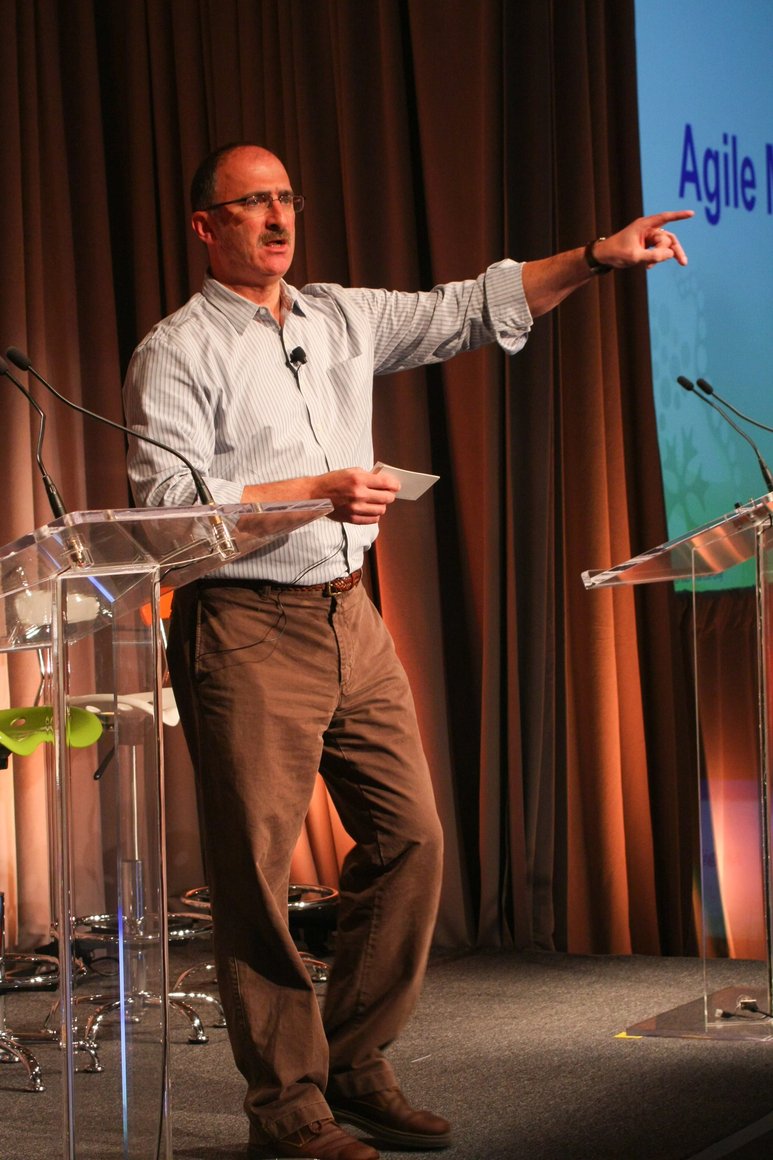 A man giving a presentation on stage, pointing at a slide that reads 'Agile.'