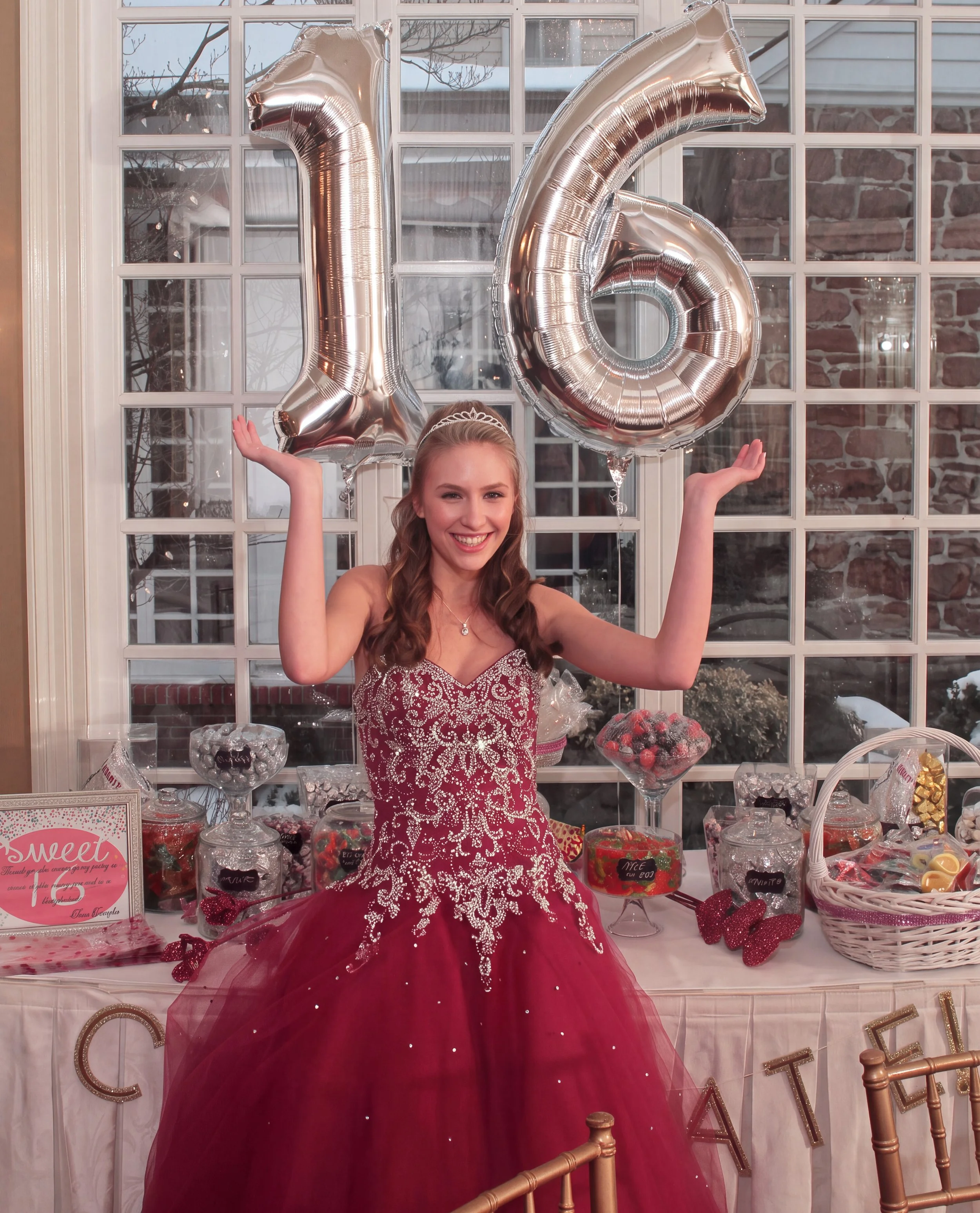 A  Sweet sixteen young woman wearing a red and white princess-style dress with a tiara, holding large silver balloons in the shape of the numbers one and six, celebrating her 16th birthday sweet 16 party in front of a decorated dessert table.