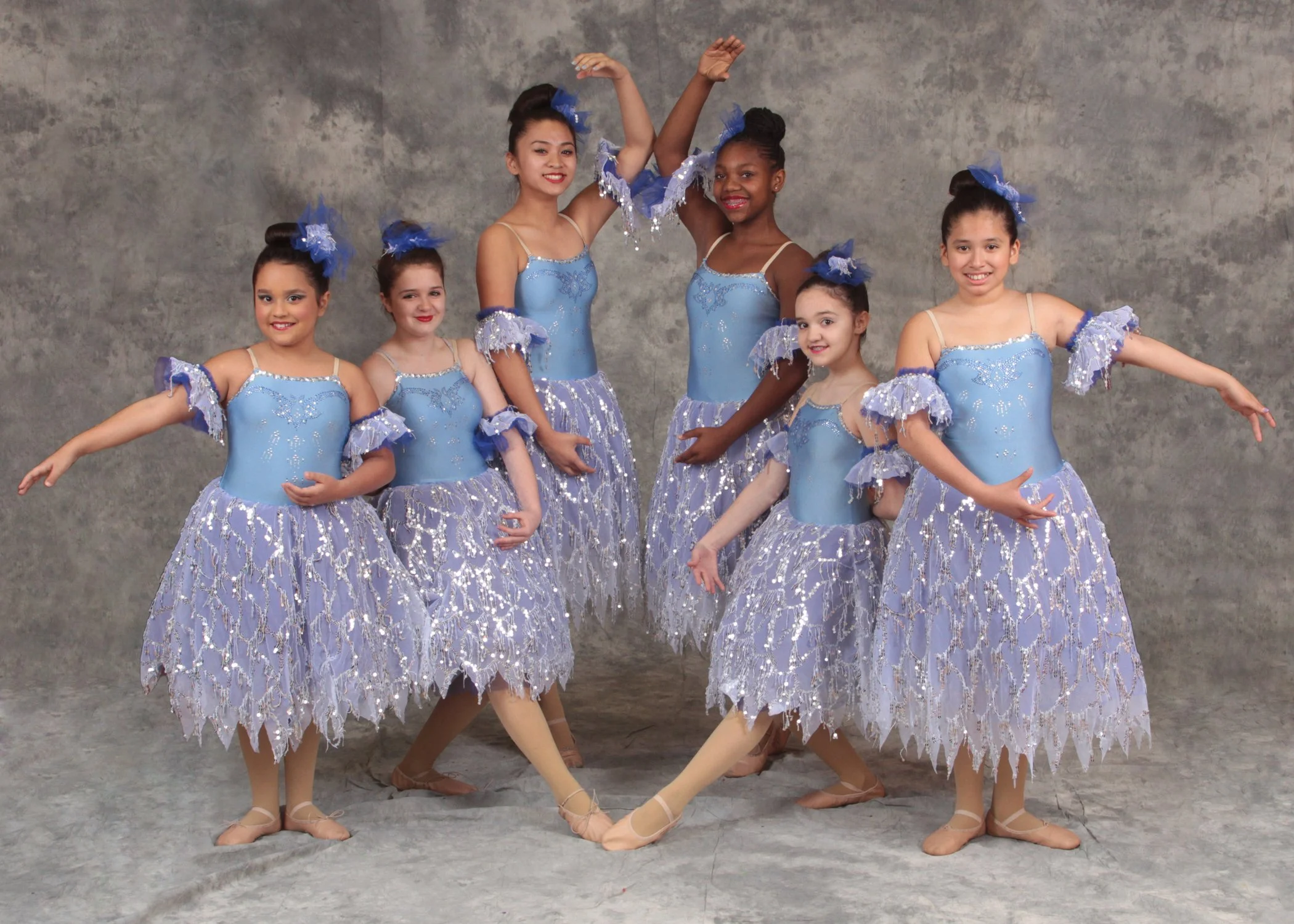 Group of seven young girls in matching blue ballet costumes with lace and sequins, posing together in front of a textured gray backdrop at a dance school in Bergen County NJ.