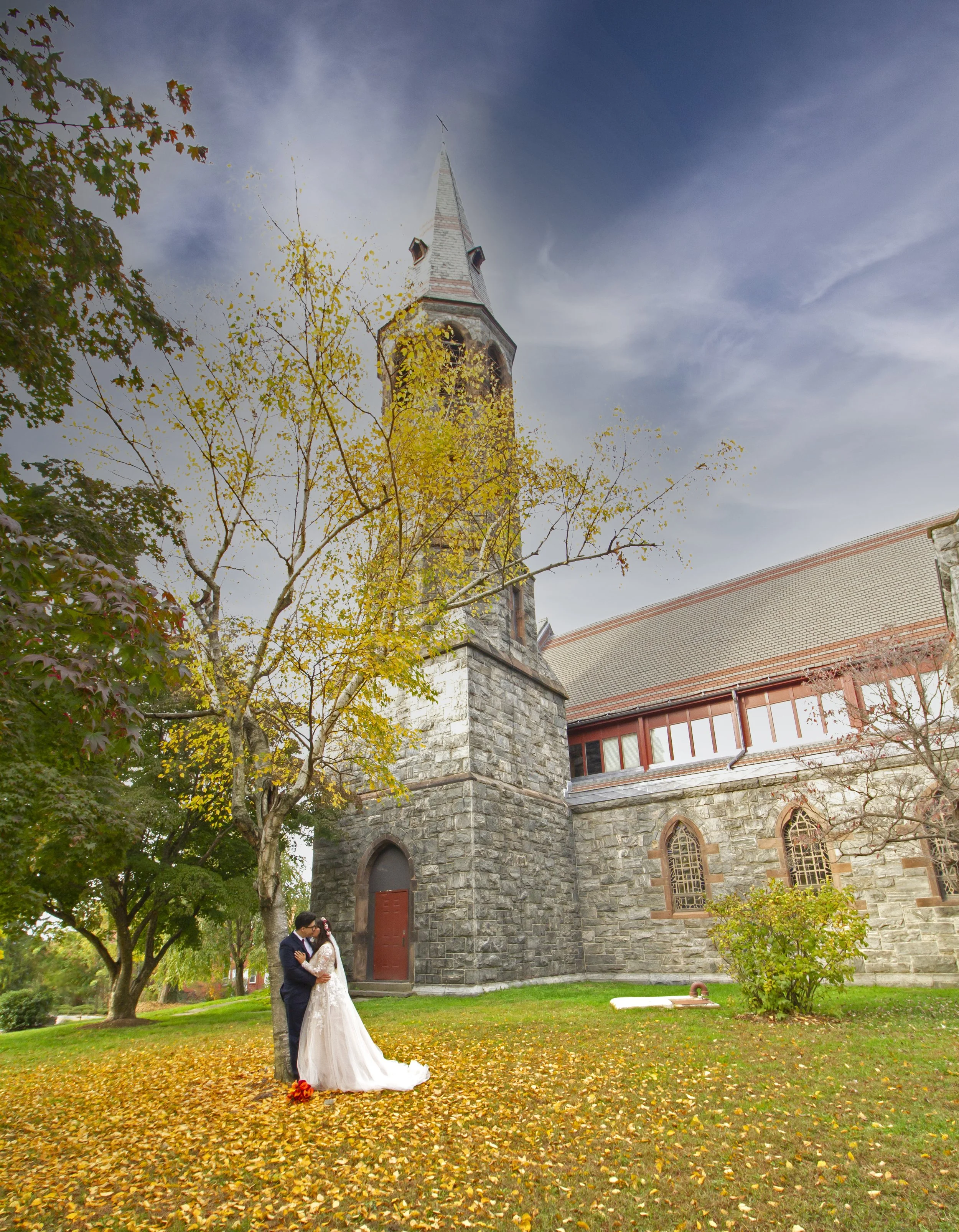 A bride and groom sharing a kiss outside a stone church during fall, with yellow and orange leaves on the ground and trees with autumn foliage in Tarrytown NY.