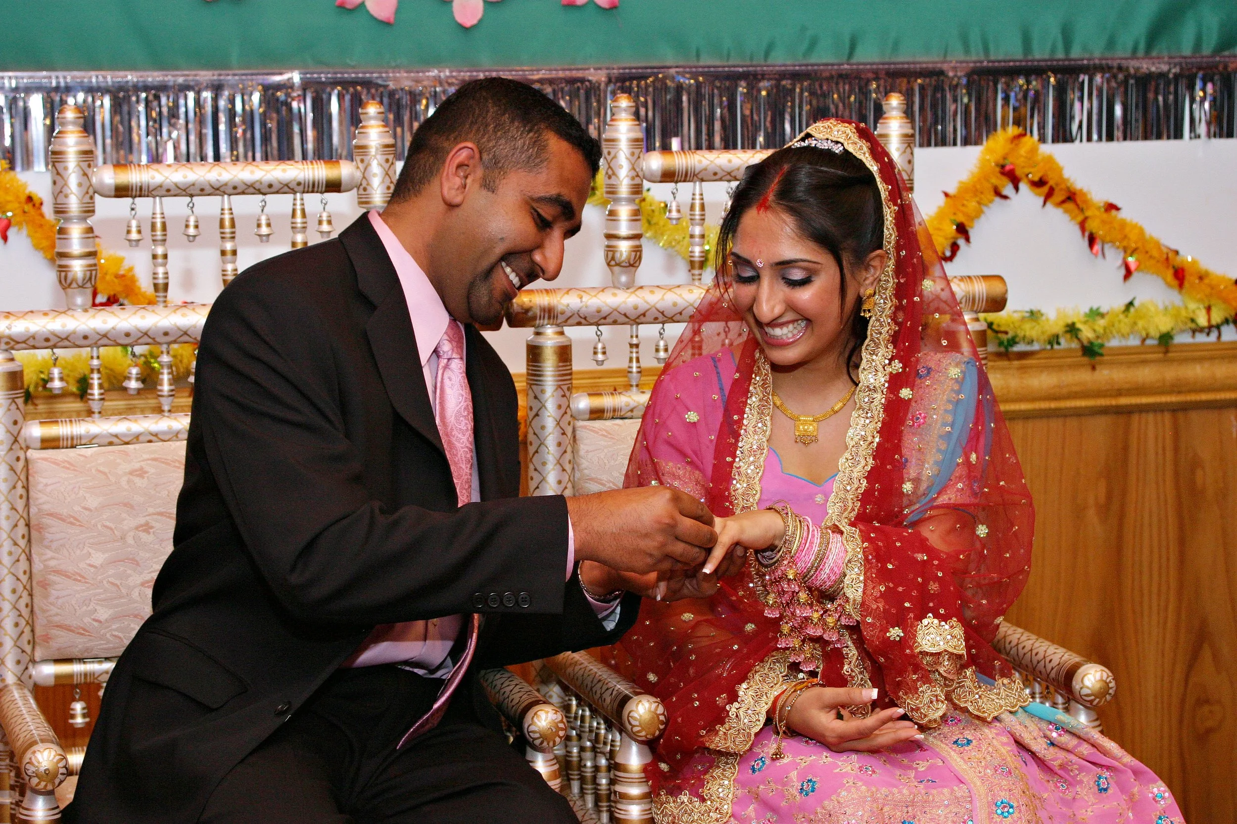 A bride and groom exchanging rings during a wedding ceremony, sitting on ornate chairs decorated with flowers, with smiling expressions.