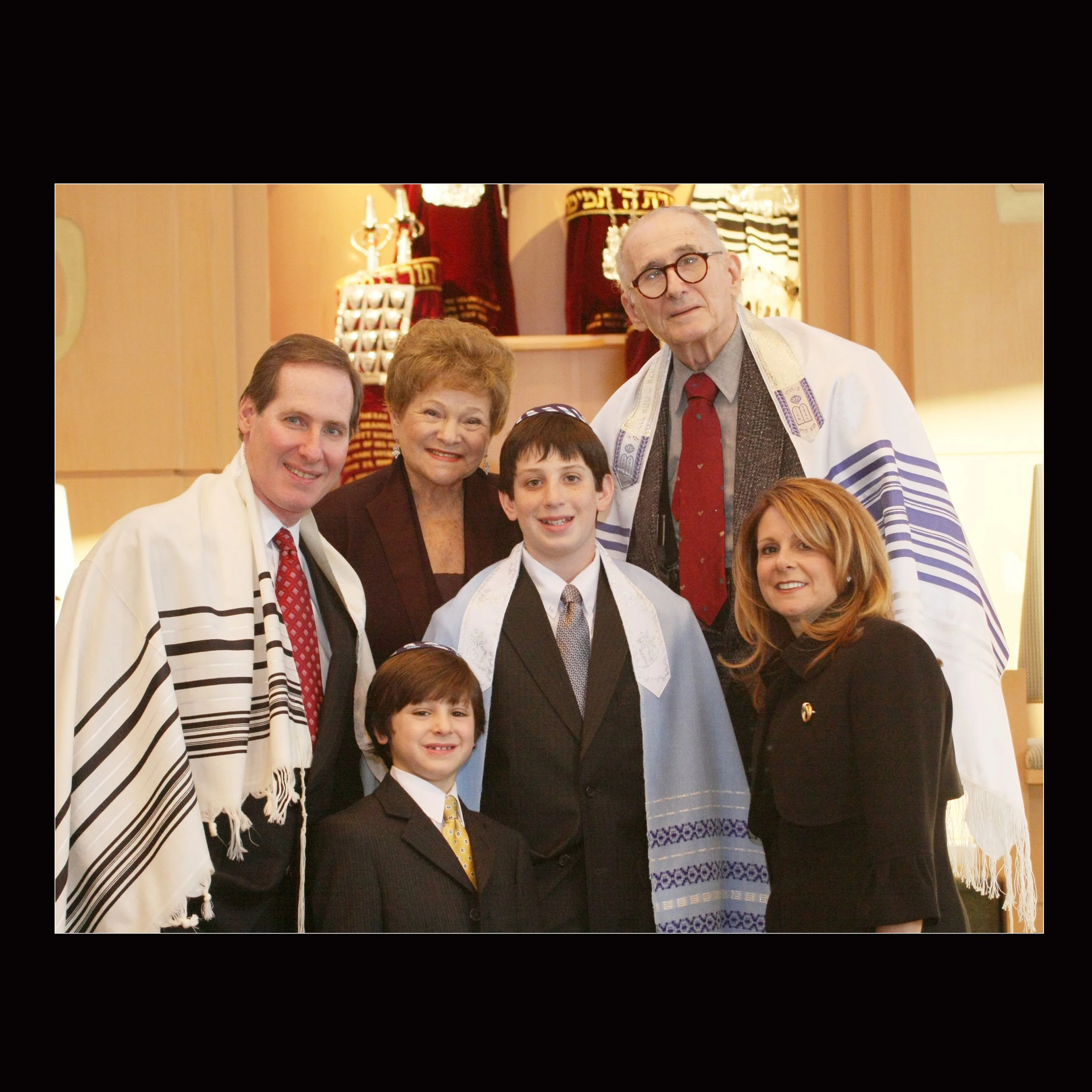 Group photo of six people, including two children, celebrating a Bar Mitzvah in a synagogue with Jewish symbols in the background.