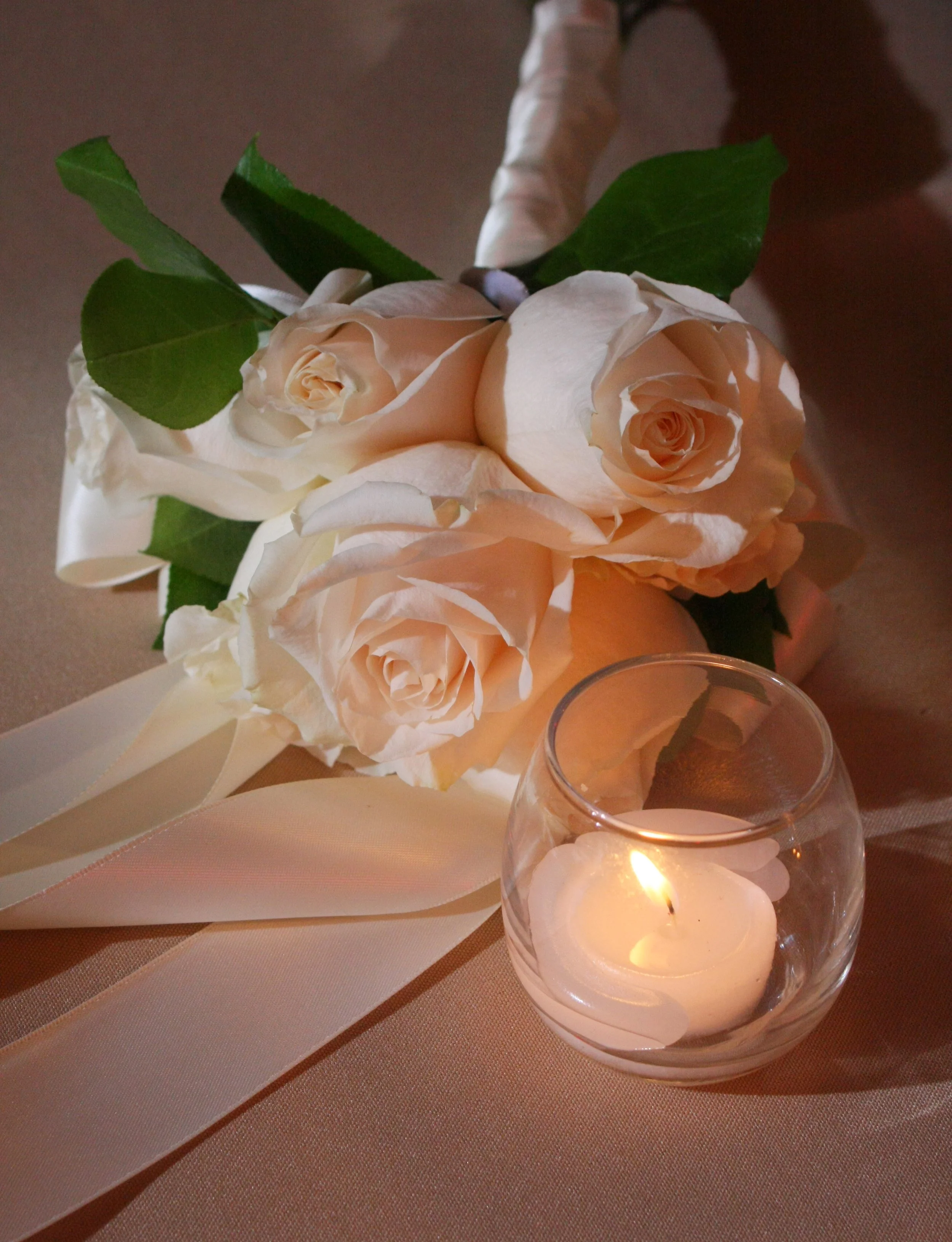 A bouquet of cream-colored roses with green leaves, tied with a white ribbon, resting on a table alongside a small lit candle in a glass holder at the Ramsey Golf and Country Club.