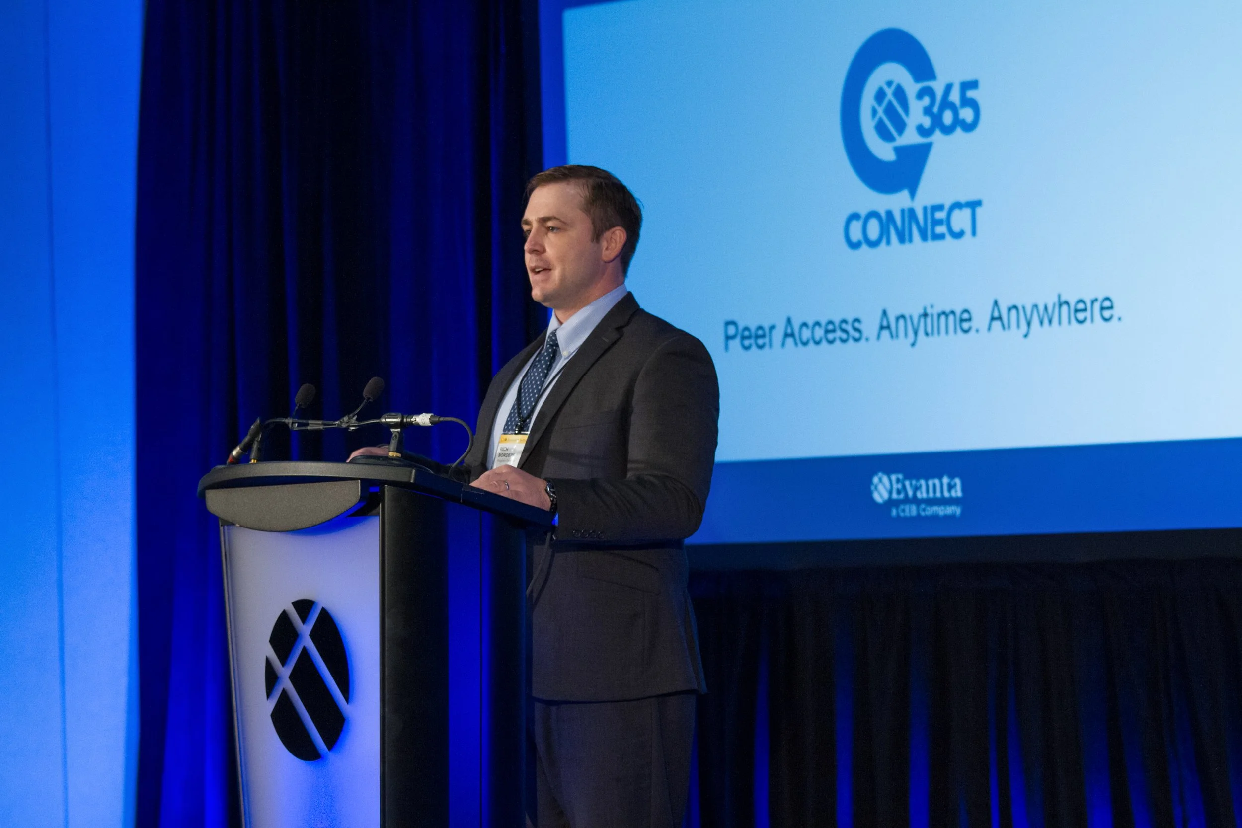 Man in a suit speaking at a conference podium with a large screen behind him displaying a presentation titled '365 Connect' with the tagline 'Peer Access. Anytime. Anywhere.'