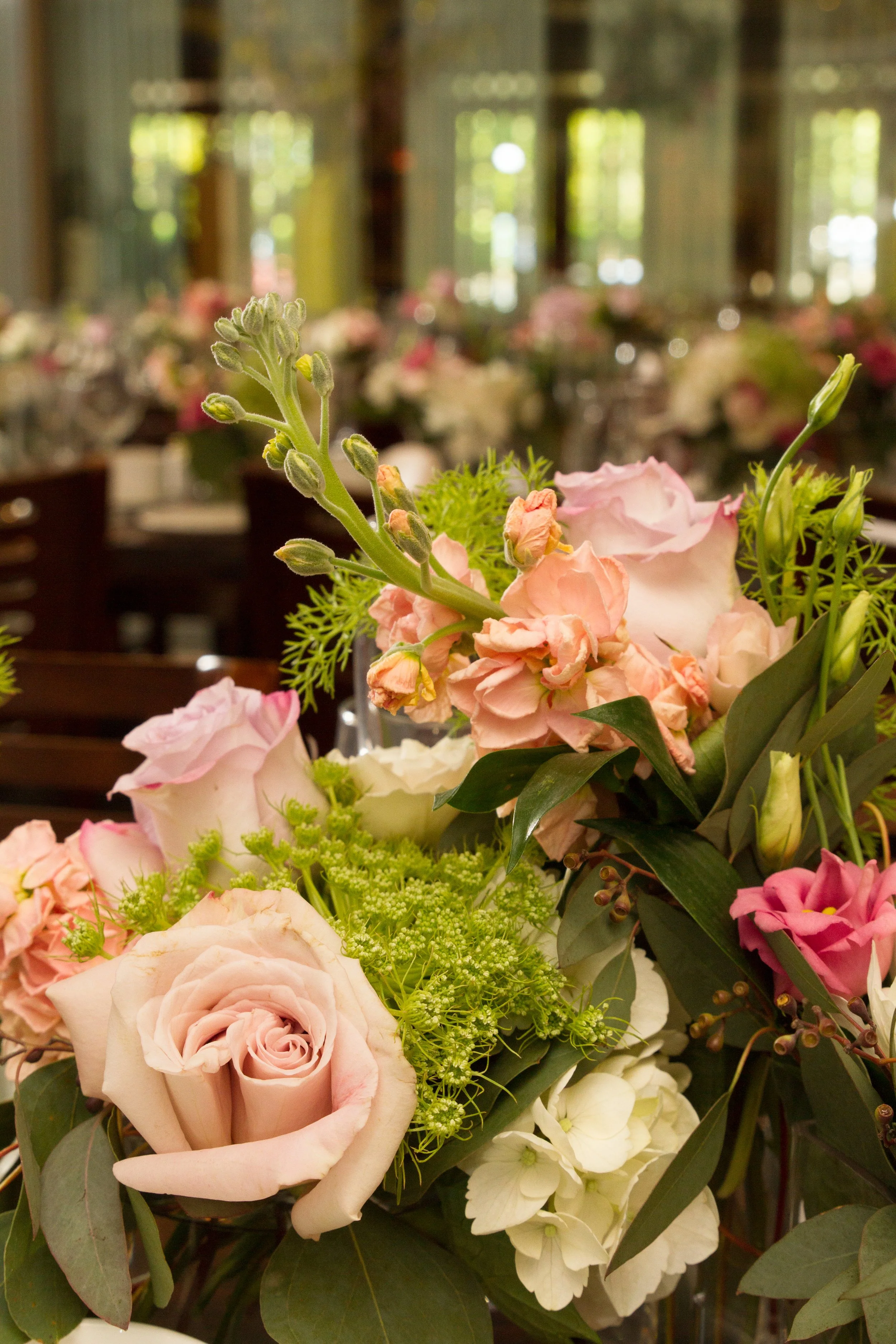 Close-up of a wedding flower arrangement with pink roses, white flowers, and greenery in a decorated indoor setting with blurred background at the Indian Trail Club.