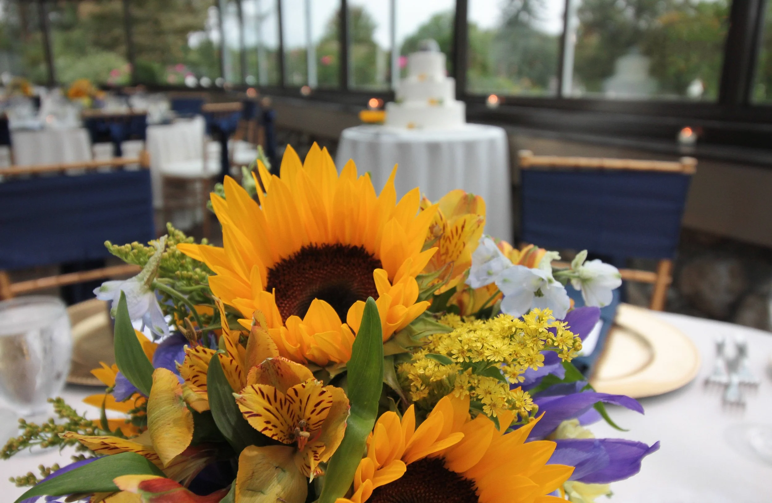 A floral centerpiece with sunflowers, yellow, purple, and white flowers on a table at an indoor event with a wedding cake in the background.