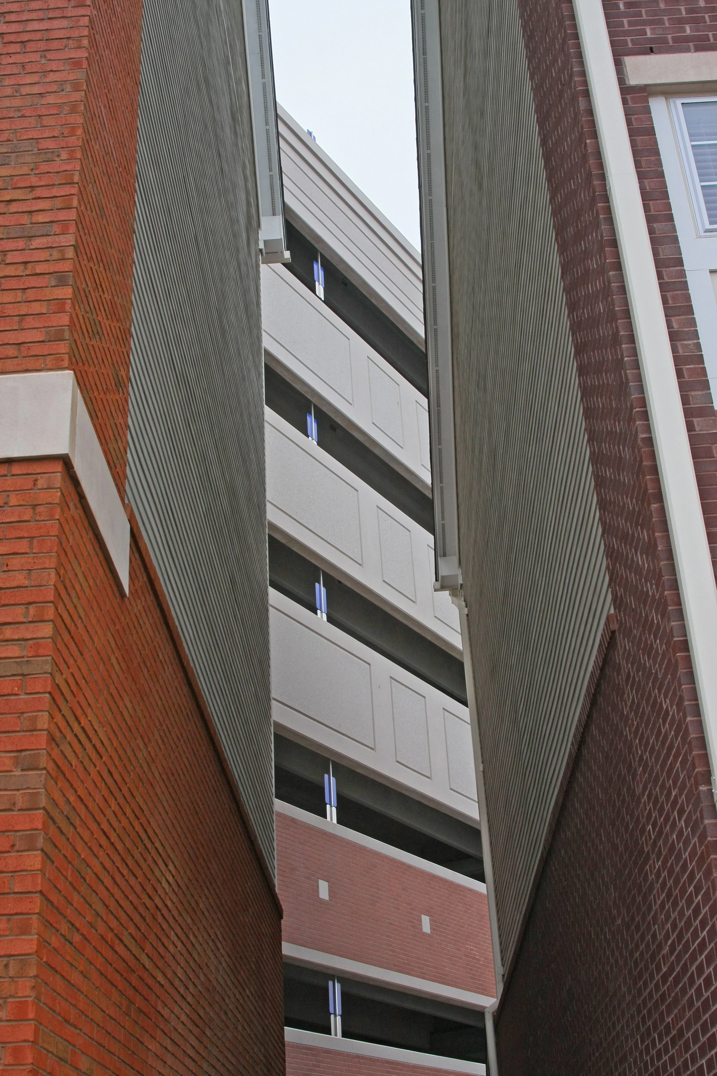 A narrow alley between a red brick building on the left and a darker brick building on the right, with a large multi-story parking garage in the background.