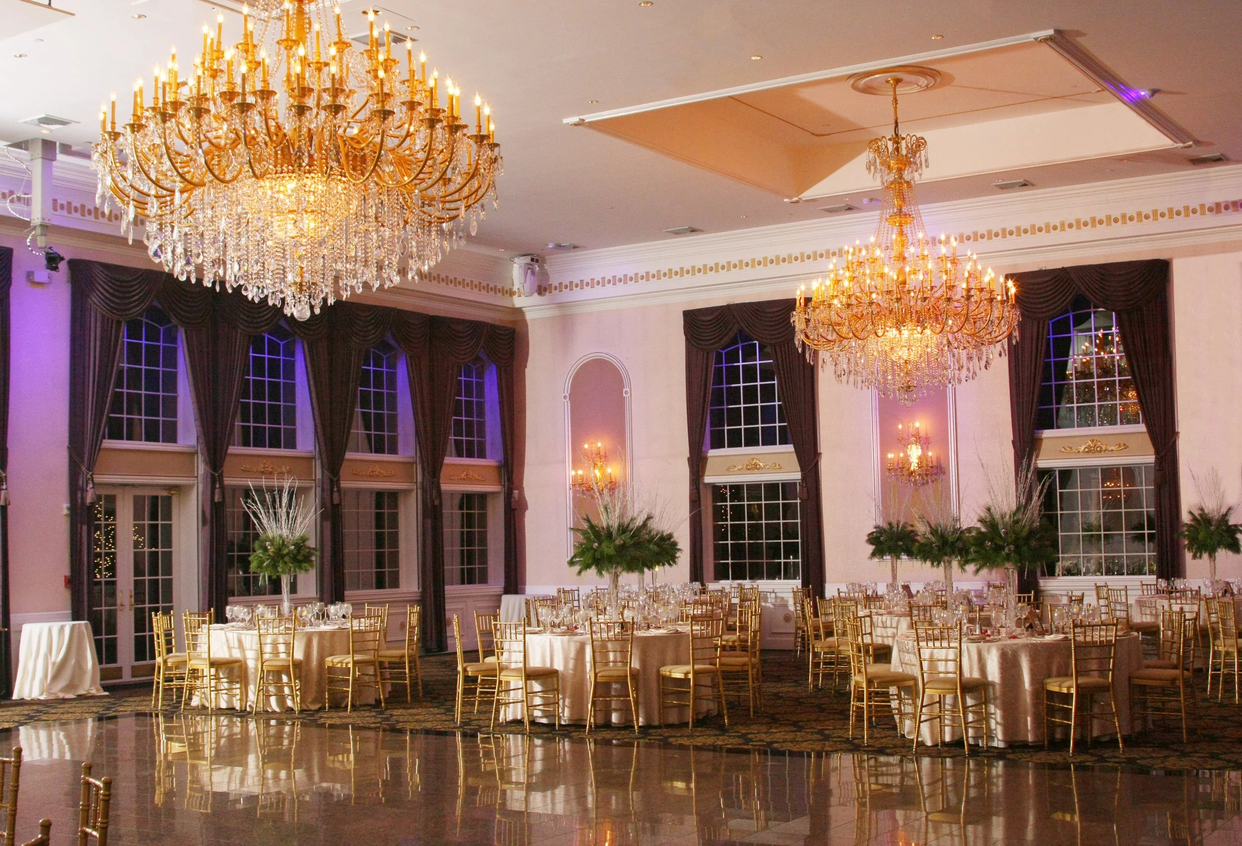 Elegant banquet hall decorated for a formal event with four large chandeliers, tall windows with dark curtains, round tables with gold chairs, and lush floral centerpieces.