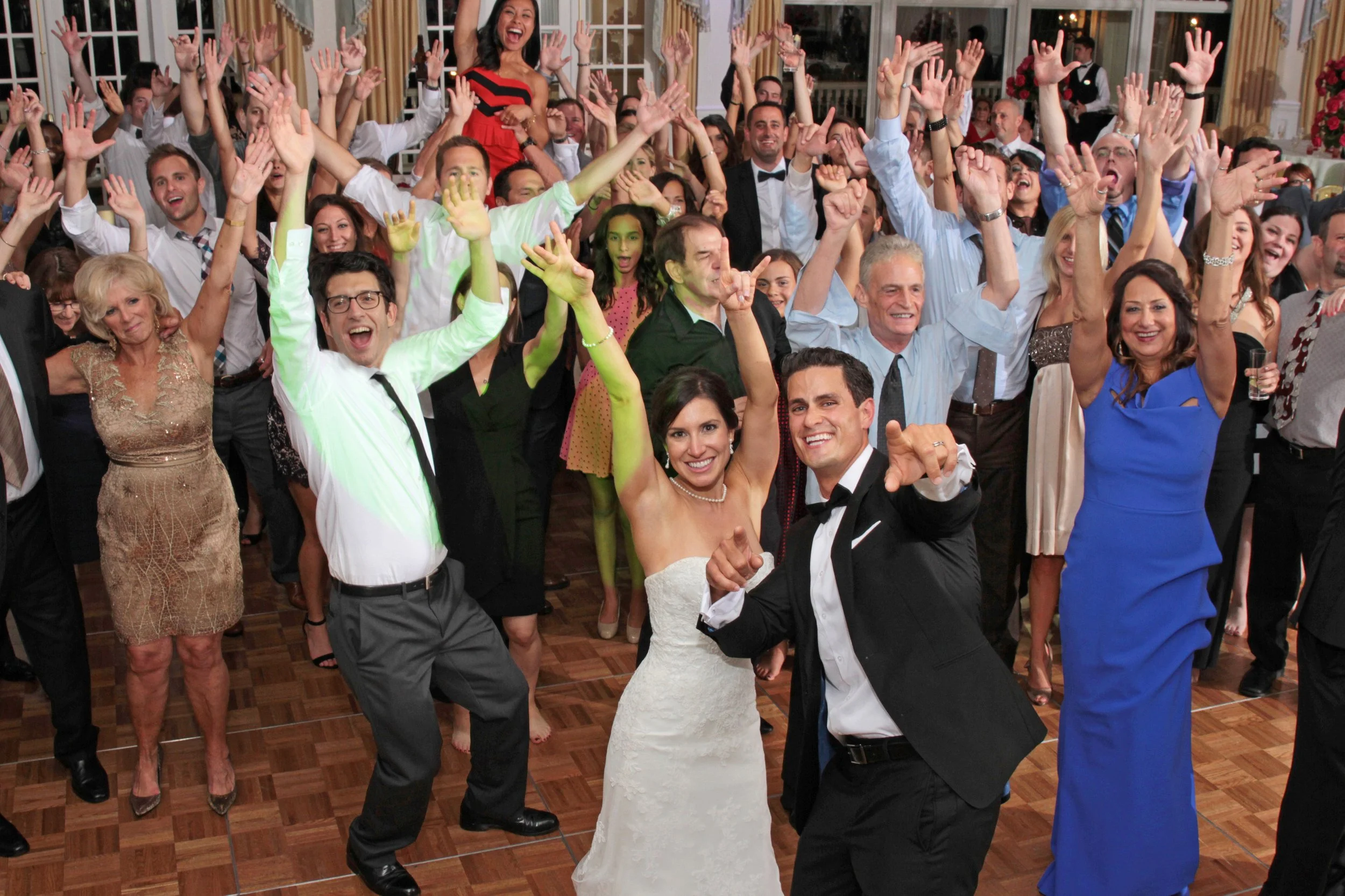 Crowd of people celebrating at a wedding reception, dancing and smiling with the bride and groom in the center.