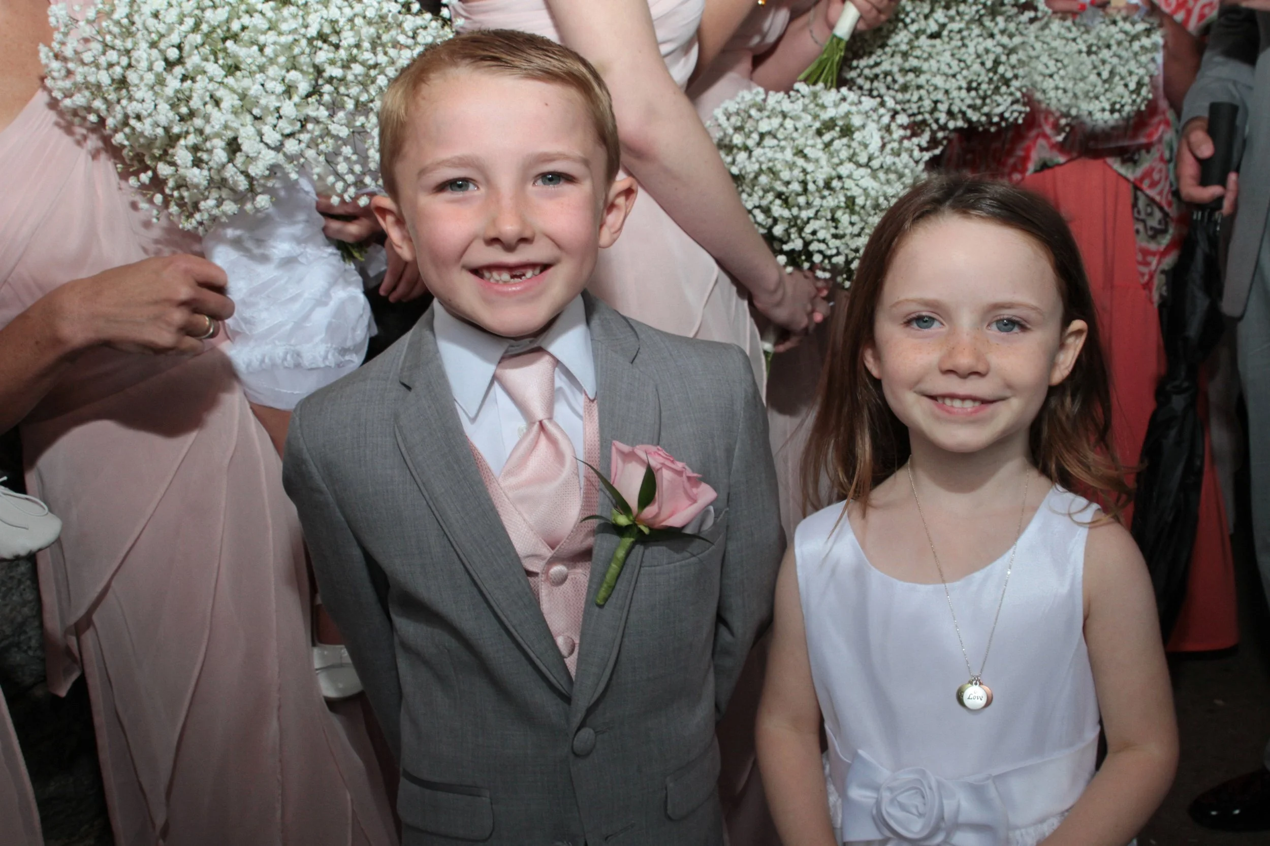 Two children dressed in formal attire smiling at a wedding or formal event, with adults holding flower bouquets in the background.
