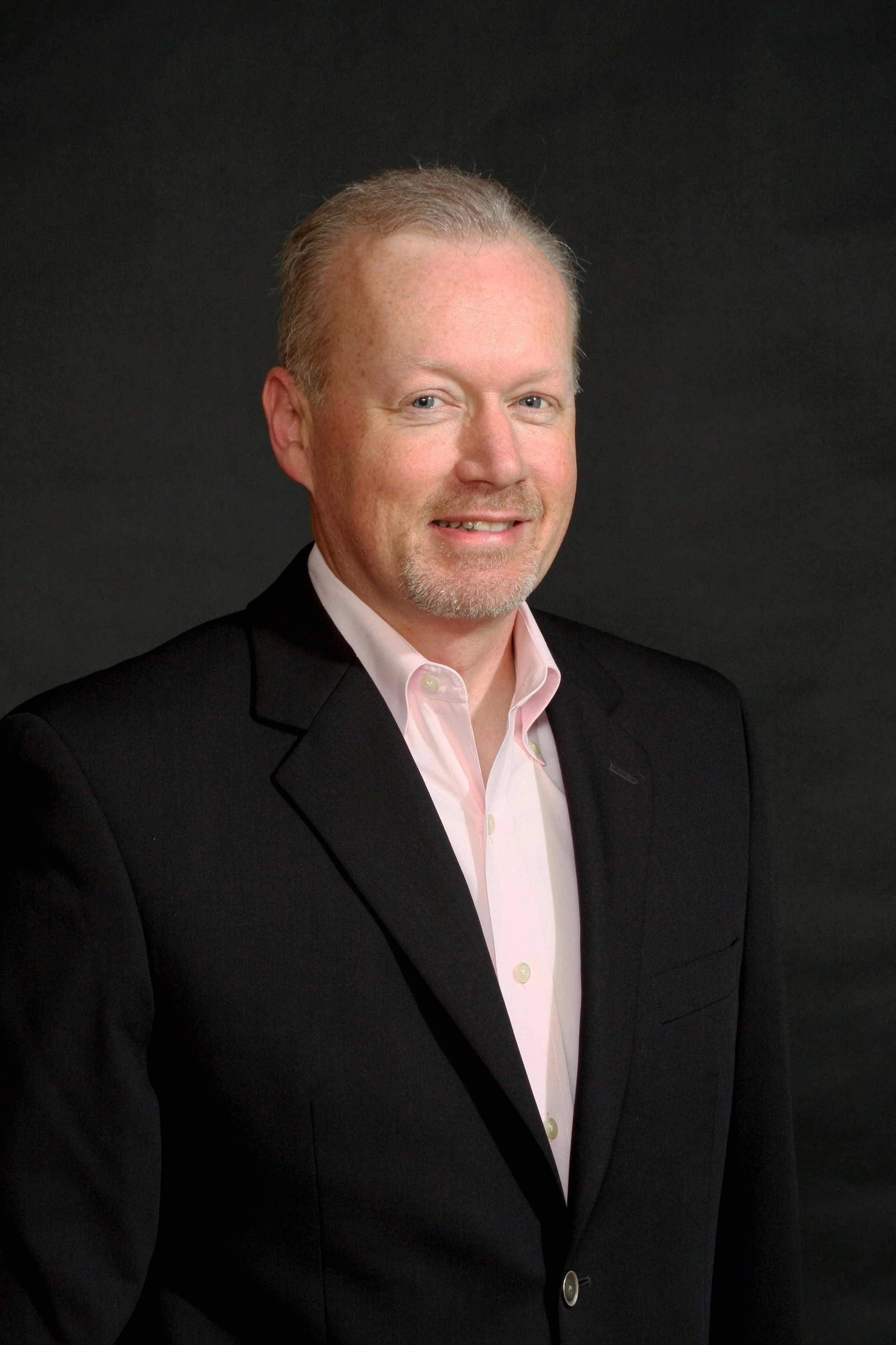 A middle-aged man with short, light-colored hair and a goatee, wearing a black suit jacket and a light pink dress shirt, smiling at the camera against a dark background.