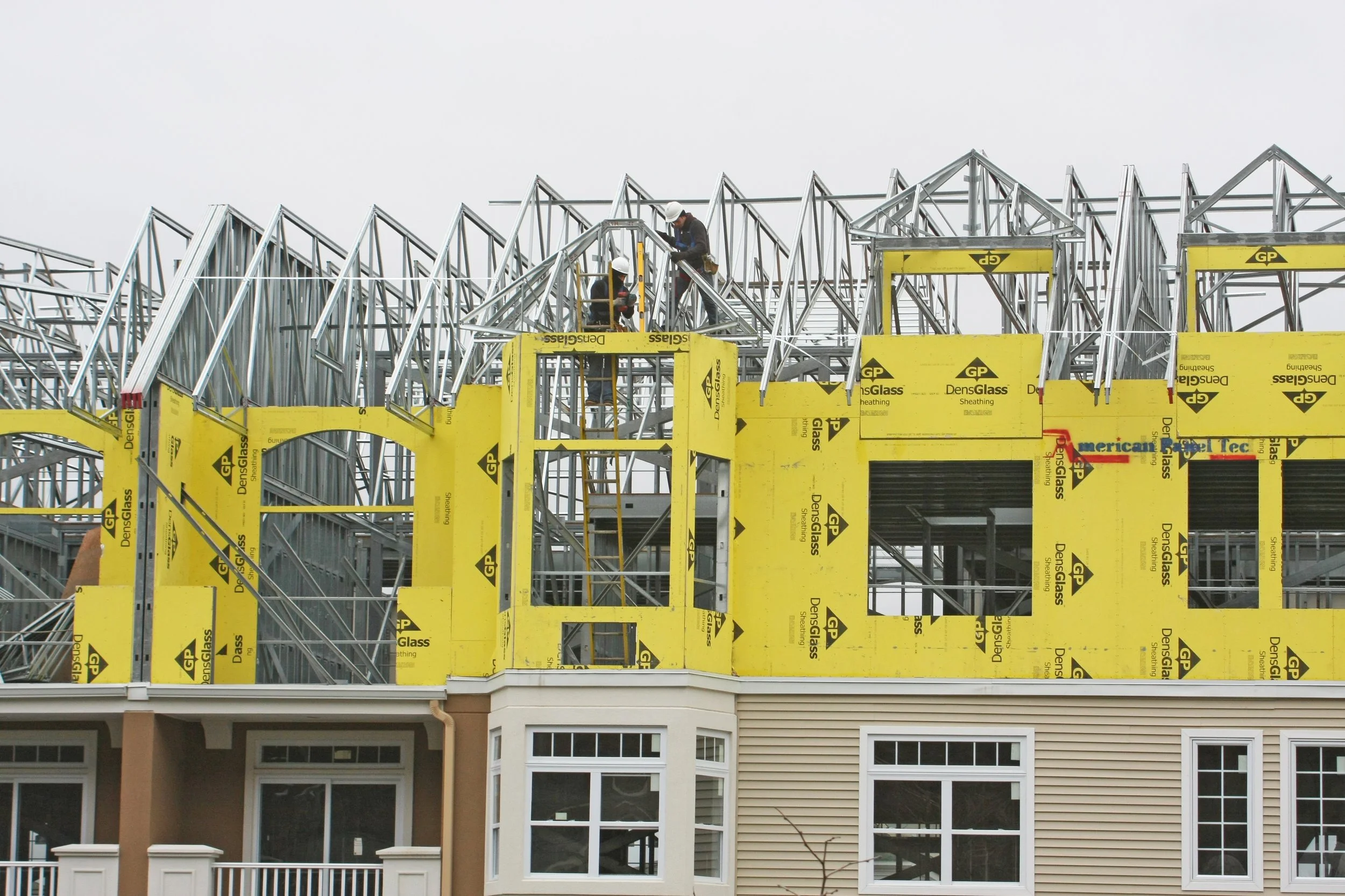 Construction workers on a partially built house frame with yellow insulation and metal framework, with a beige and white building below.