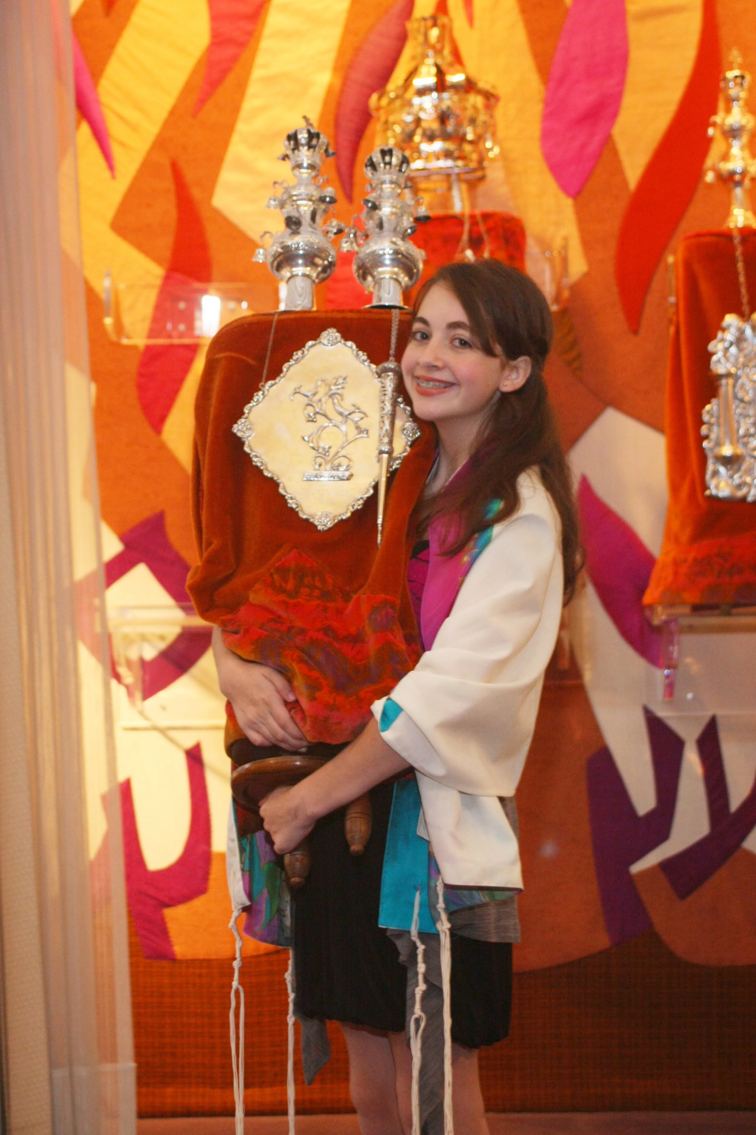 A young Mitzvah woman with long brown hair holding a large, ornate, red velvet torah with silver embellishments, smiling in a temple with colorful, patterned artwork in the background.