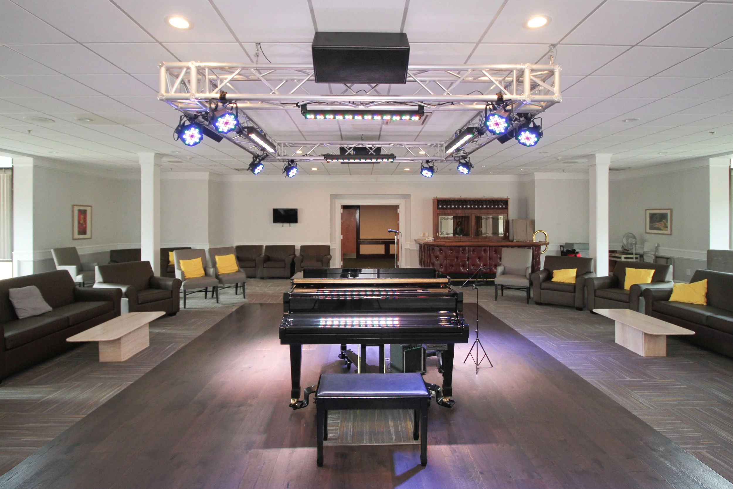 An indoor lounge with a central black grand piano, surrounded by black sofas with yellow cushions, wooden benches, and a bar in the background. Overhead stage lighting is mounted on a truss.