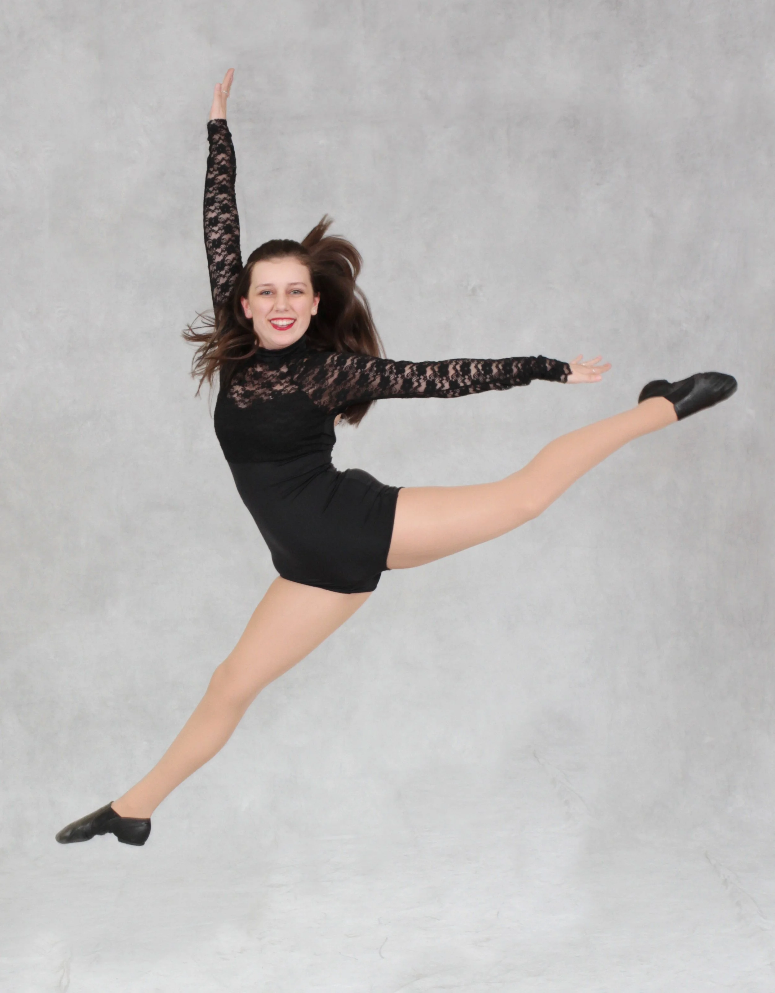 A woman jumping in the air wearing a black lace top, black shorts, beige tights, and black shoes, smiling at the camera with a gray textured background at a dance school in Bergen County NJ.