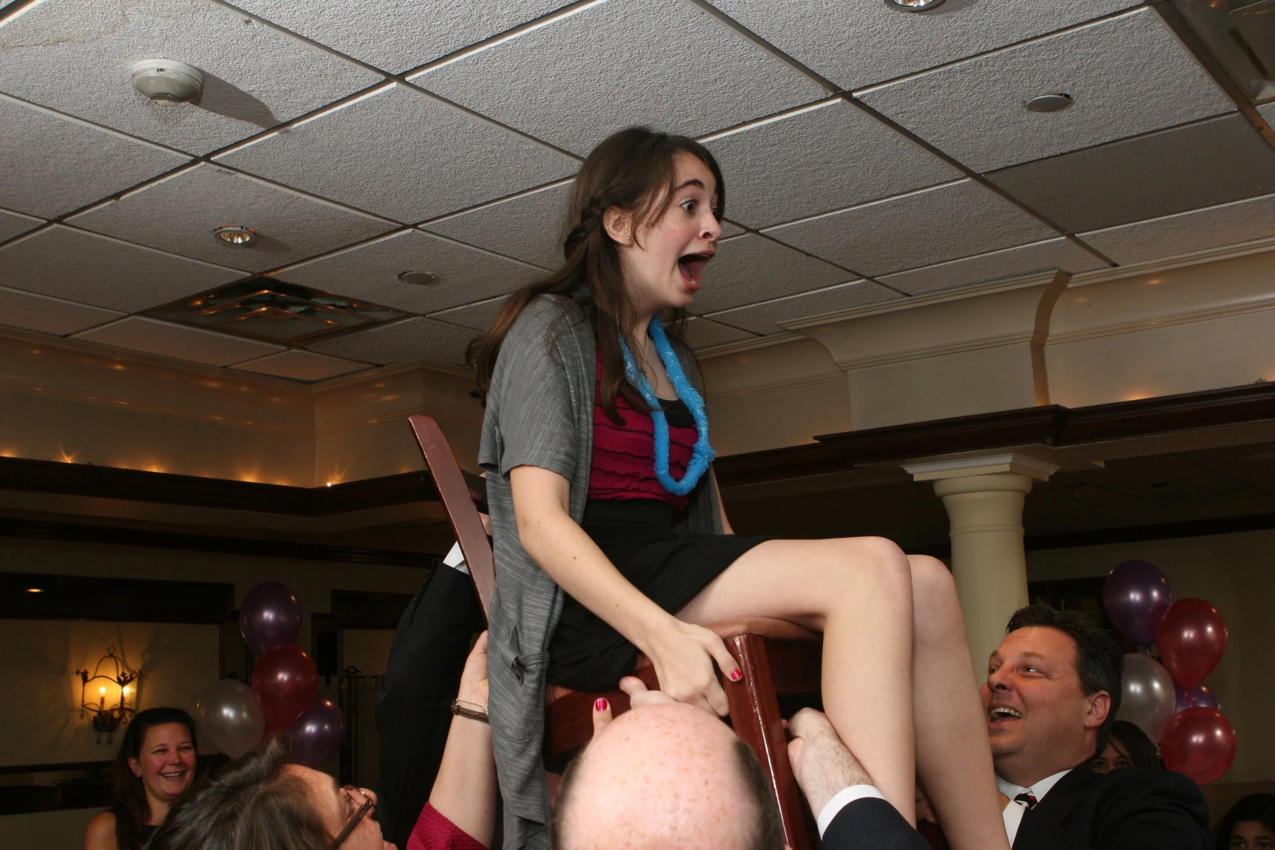 A young Bat Mitzvah girl sitting on a chair being lifted by people at a hooray celebration event, with surprised and excited expressions, and balloons in the background.