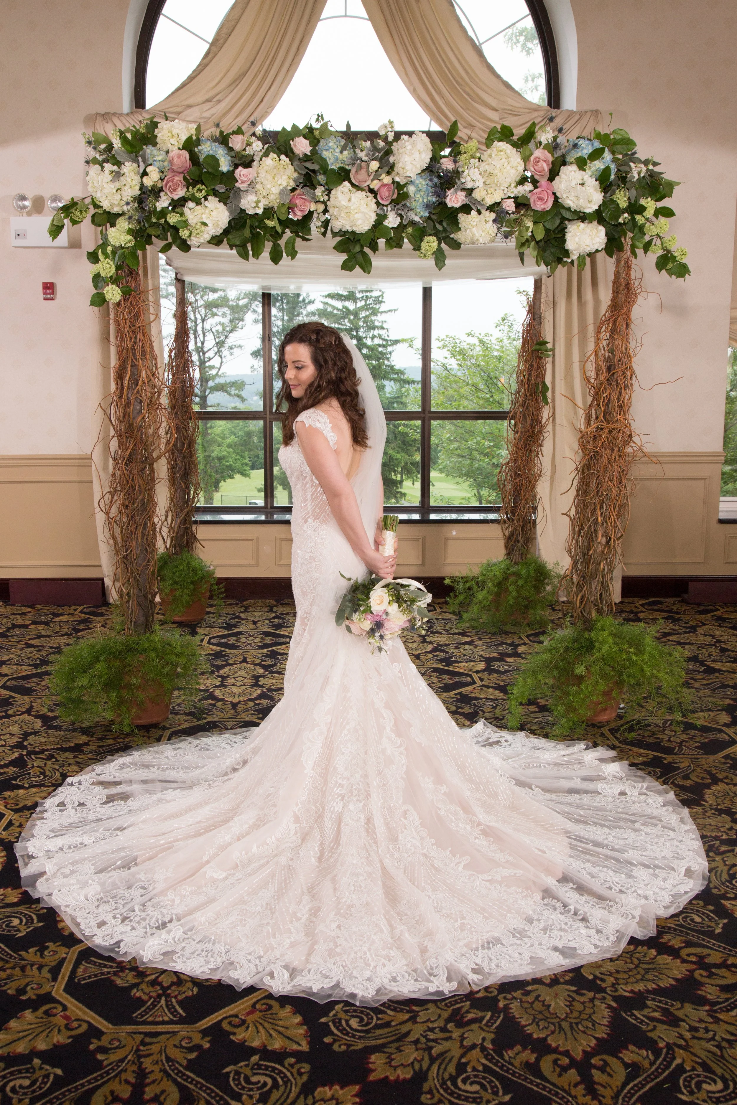 A bride in a white lace wedding dress holding a bouquet stands on a patterned carpet in front of a floral wedding arch with greenery, pink and white flowers, and wood accents.