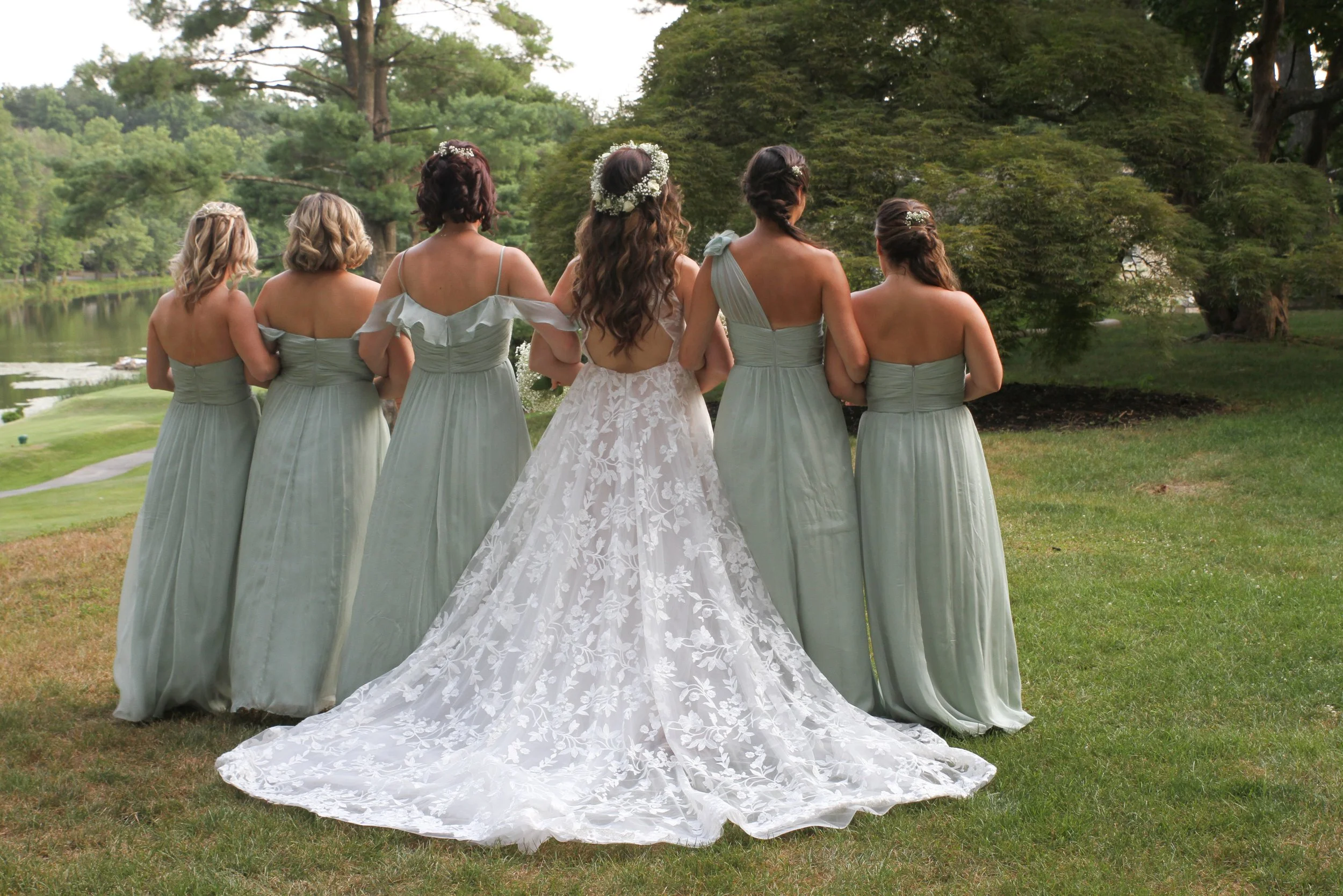 Bride in a white lace wedding gown with a train walking with six bridesmaids in matching light green dresses on a grassy area near a pond with trees.