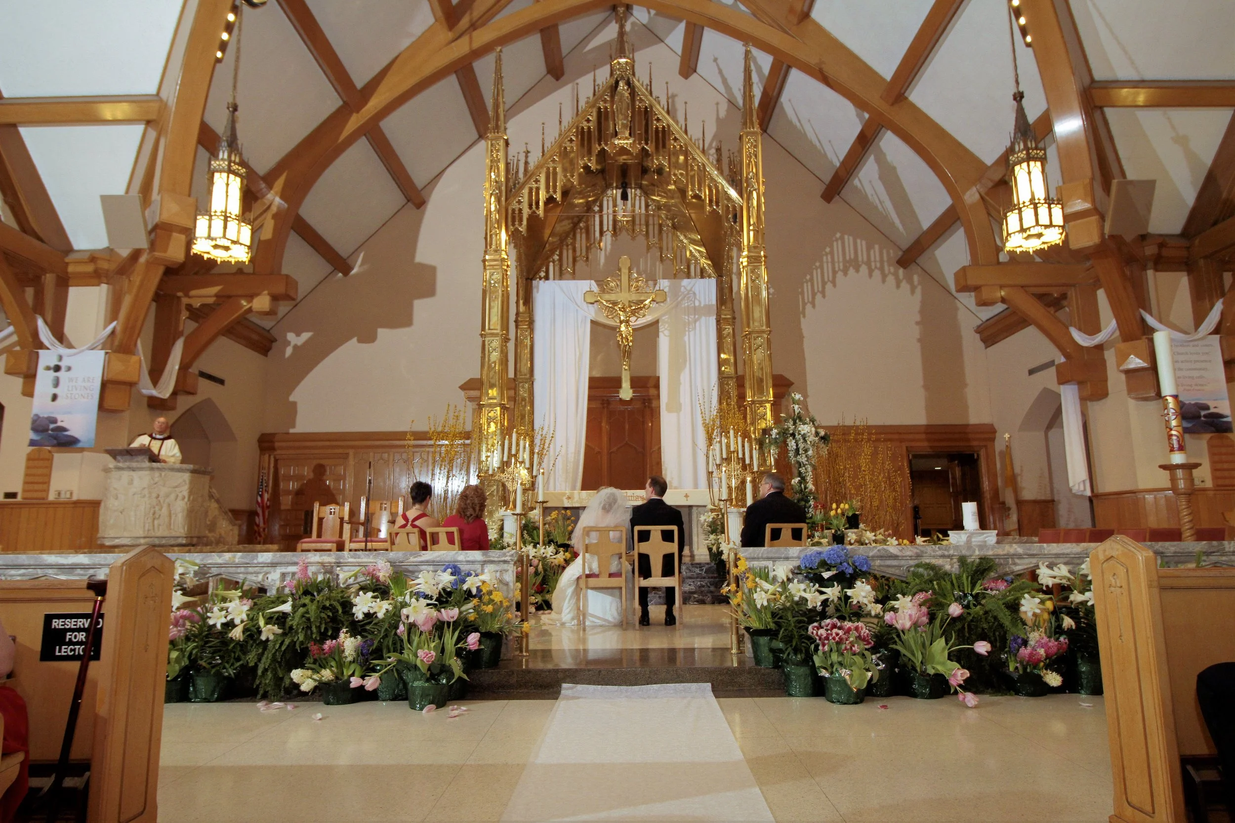 Interior of a church decorated for a wedding, with a bride and groom sitting at the altar facing the officiant. The church has wooden beams, gold accents, and floral arrangements along the aisle.