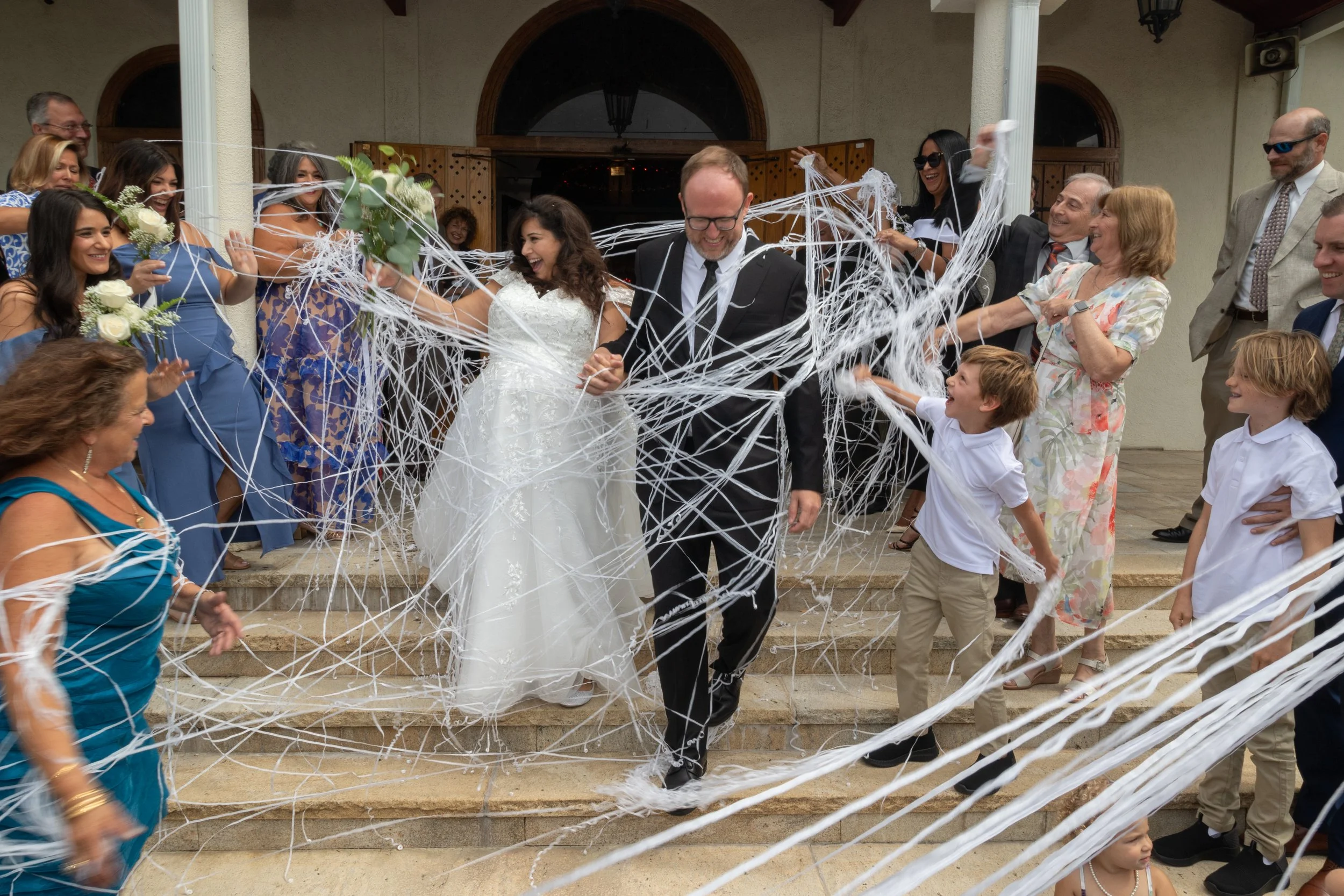 Wedding celebration with a bride, groom, and guests releasing white streamers or confetti on steps outside a building.