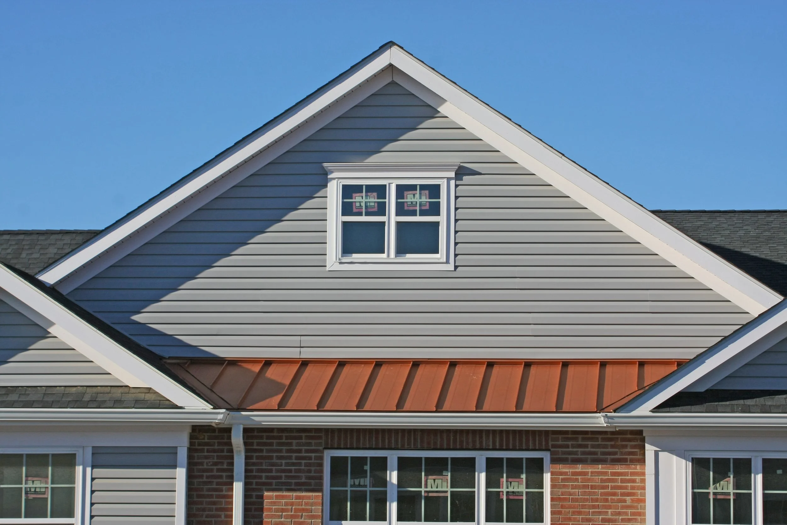 Close-up view of a house roof with gray vinyl siding, a small window, and a partially visible red metal roof in bright sunlight under a clear blue sky.