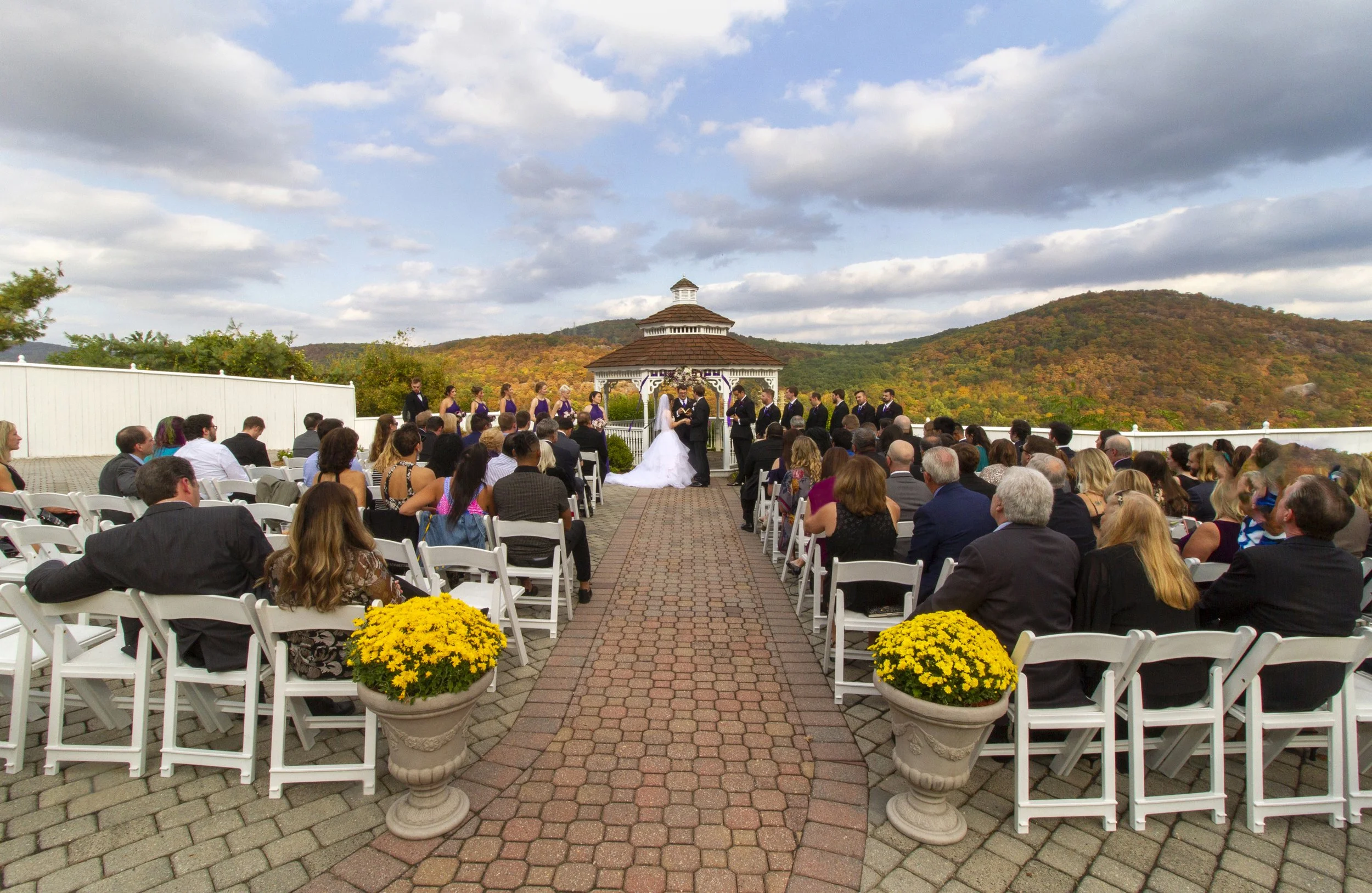 An outdoor wedding ceremony with guests seated on white chairs, a bride and groom at the altar, and a scenic mountain backdrop with cloudy sky at the Garrison.
