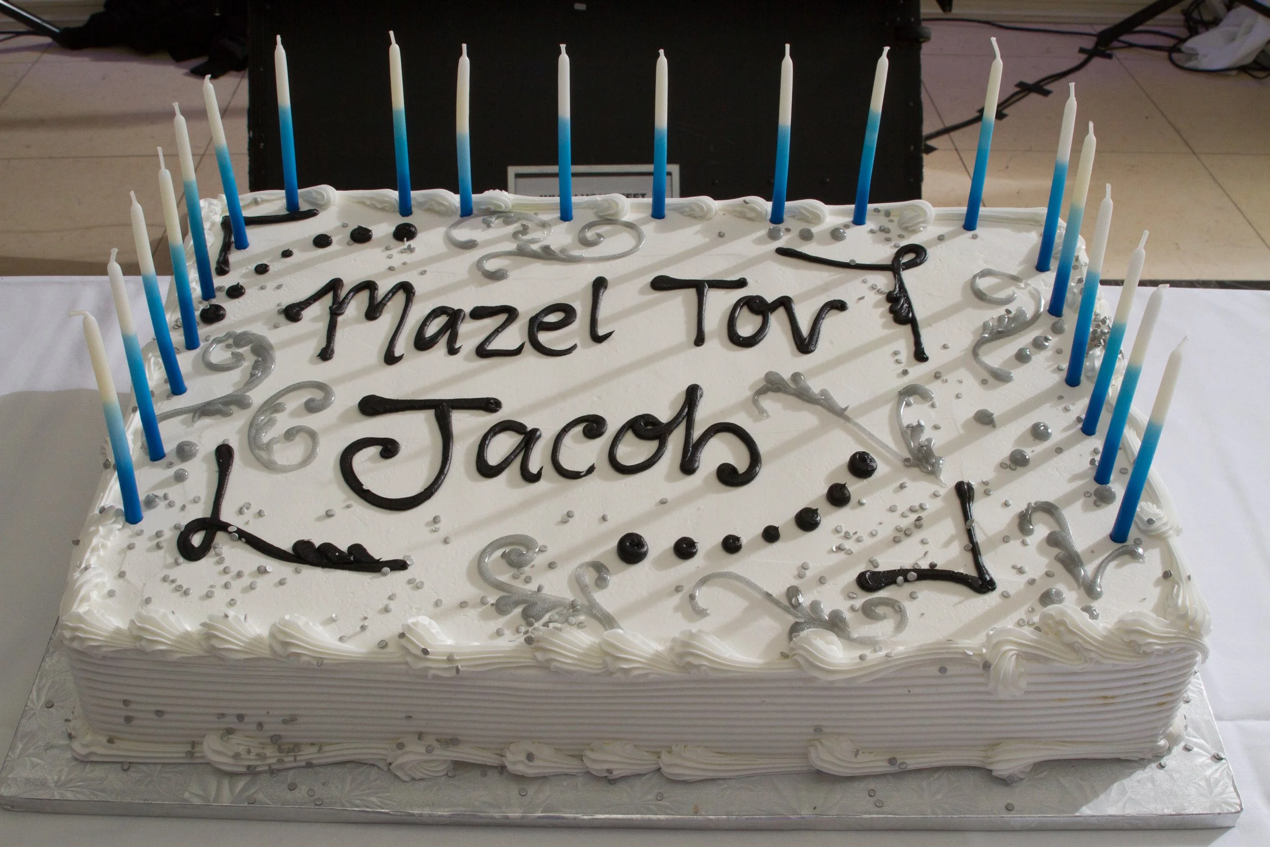 A large rectangular Bar Mitzvah cake with white frosting, decorated with black and silver icing, and blue candles along the edges. The cake has the message 'Mazel Tov Jacob' written on top.