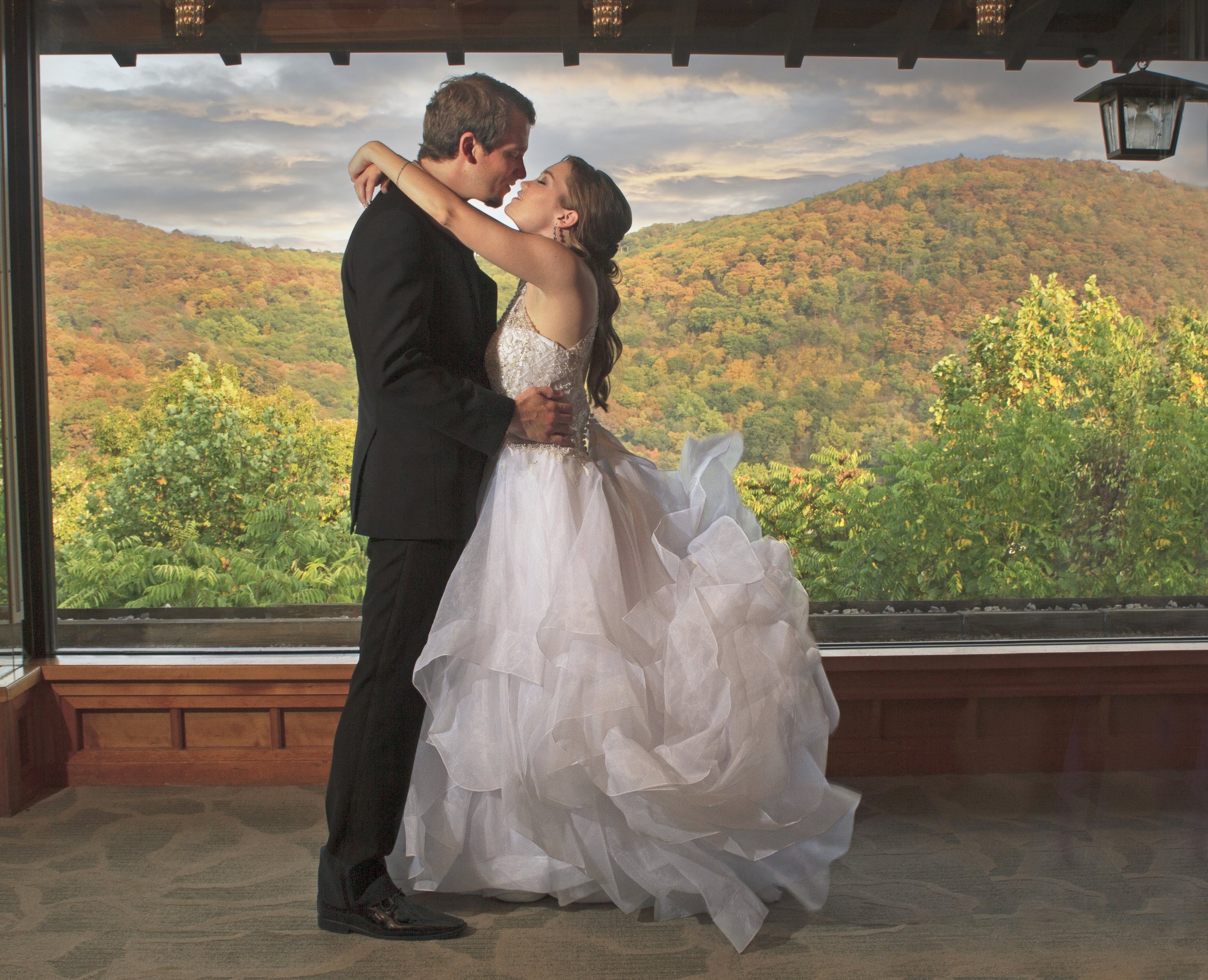 A bride and groom at the Views at Mount Fuji dancing close together indoors with a large window overlooking a mountain landscape with colorful autumn trees.