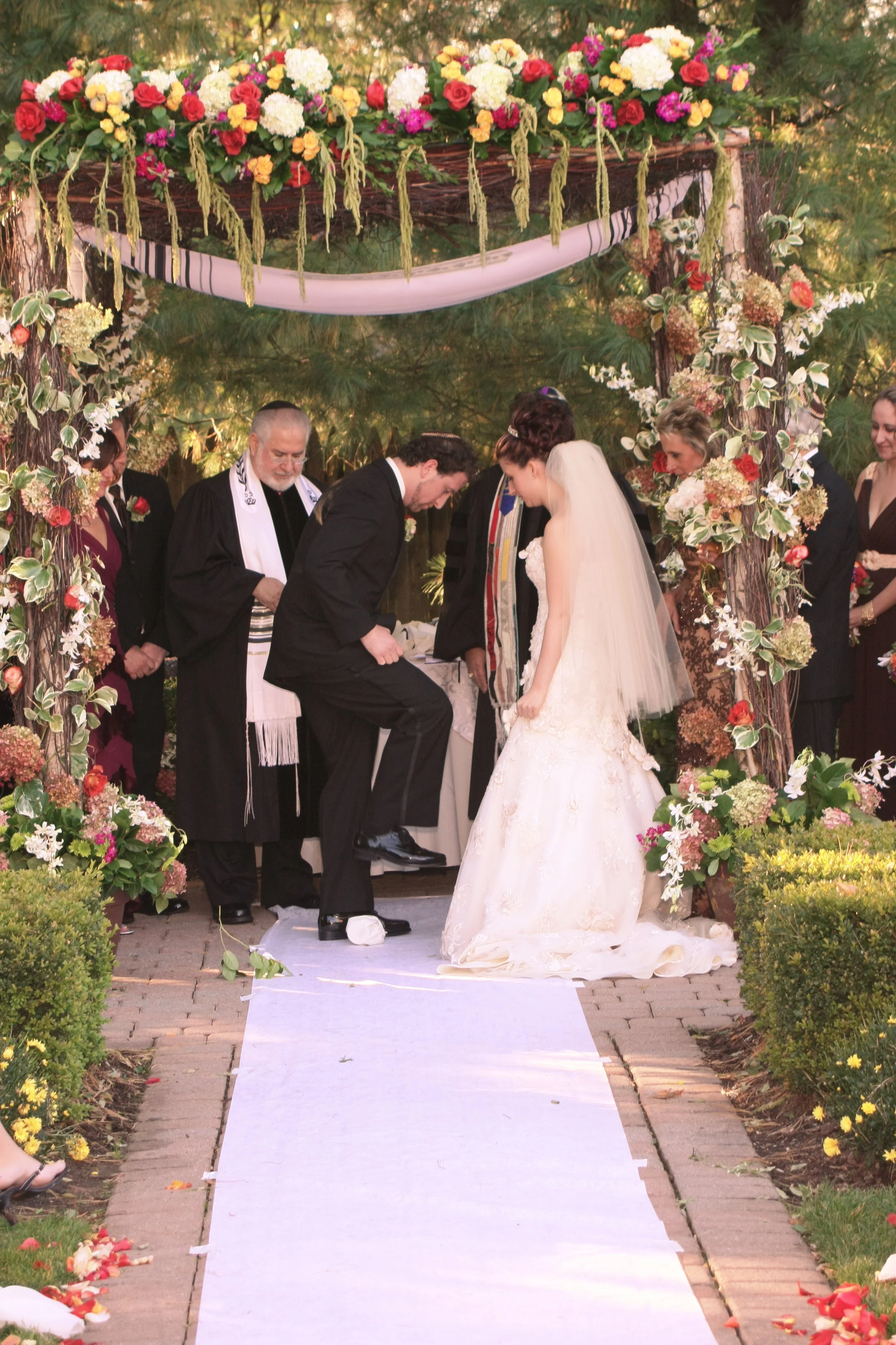 A wedding ceremony outdoors with a bride and groom exchanging vows under a floral arch, with officiants and guests present.