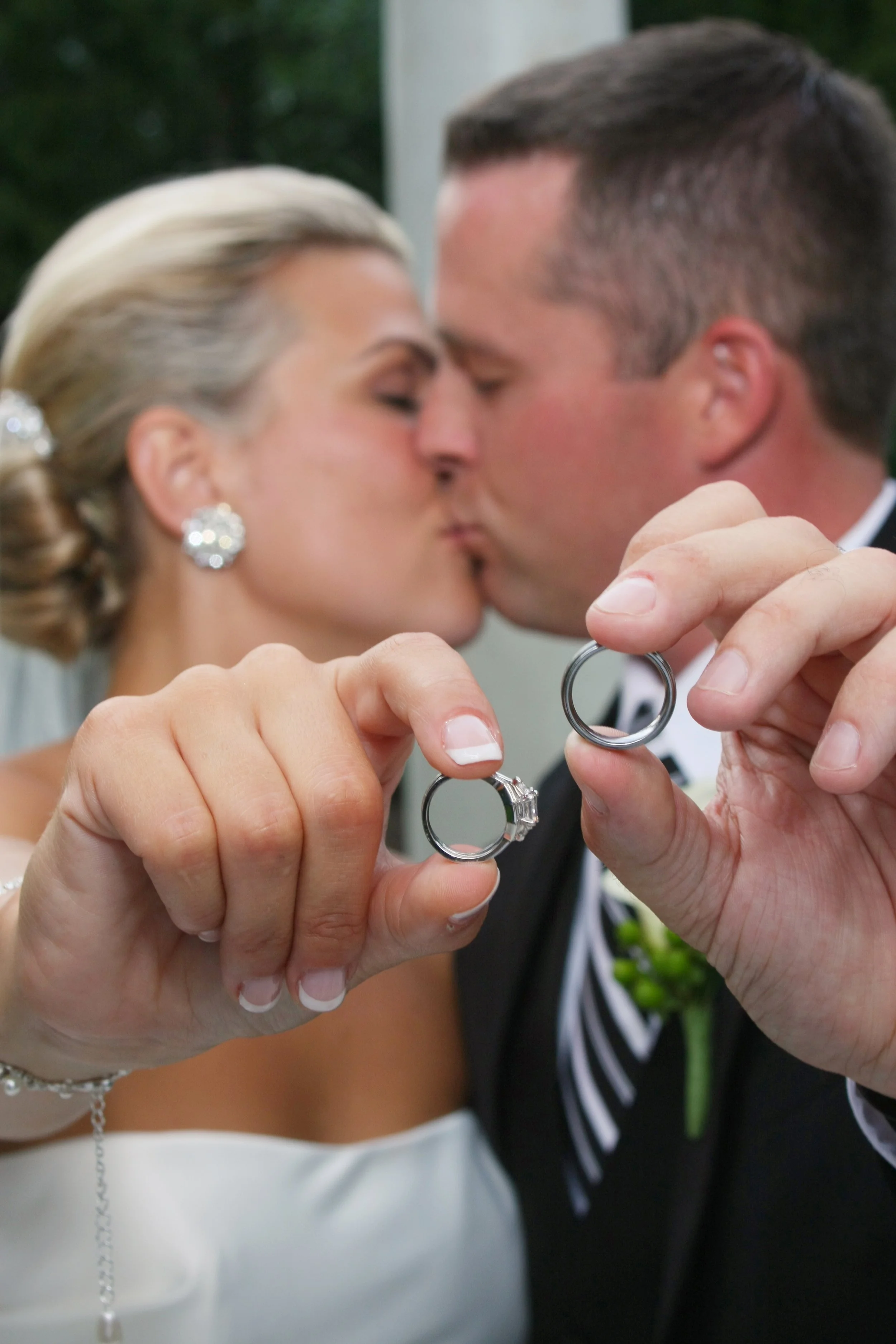 A bride and groom sharing a kiss while holding up wedding rings in front of them.