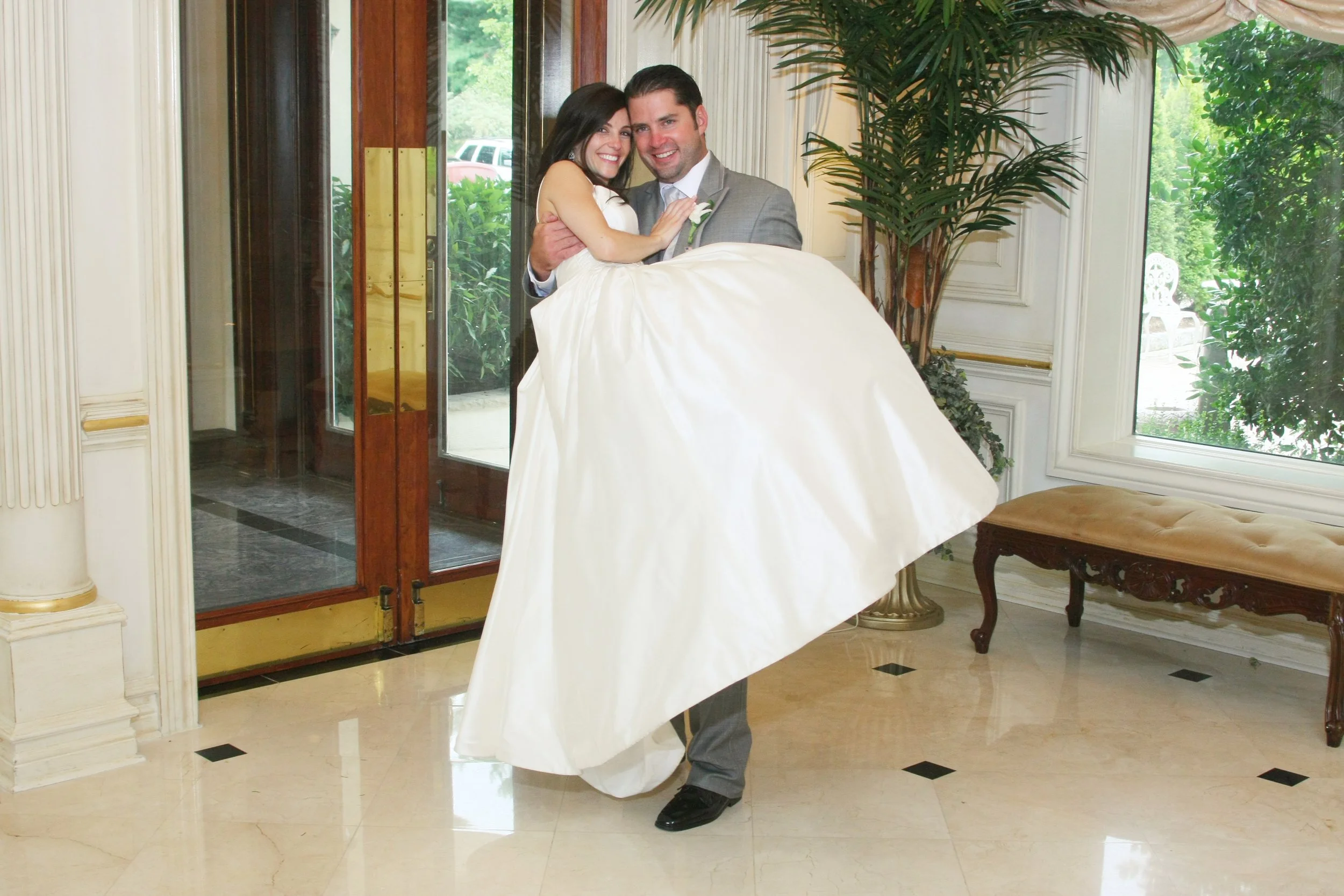 Groom lifting bride in a wedding dress indoors with large windows and plants.