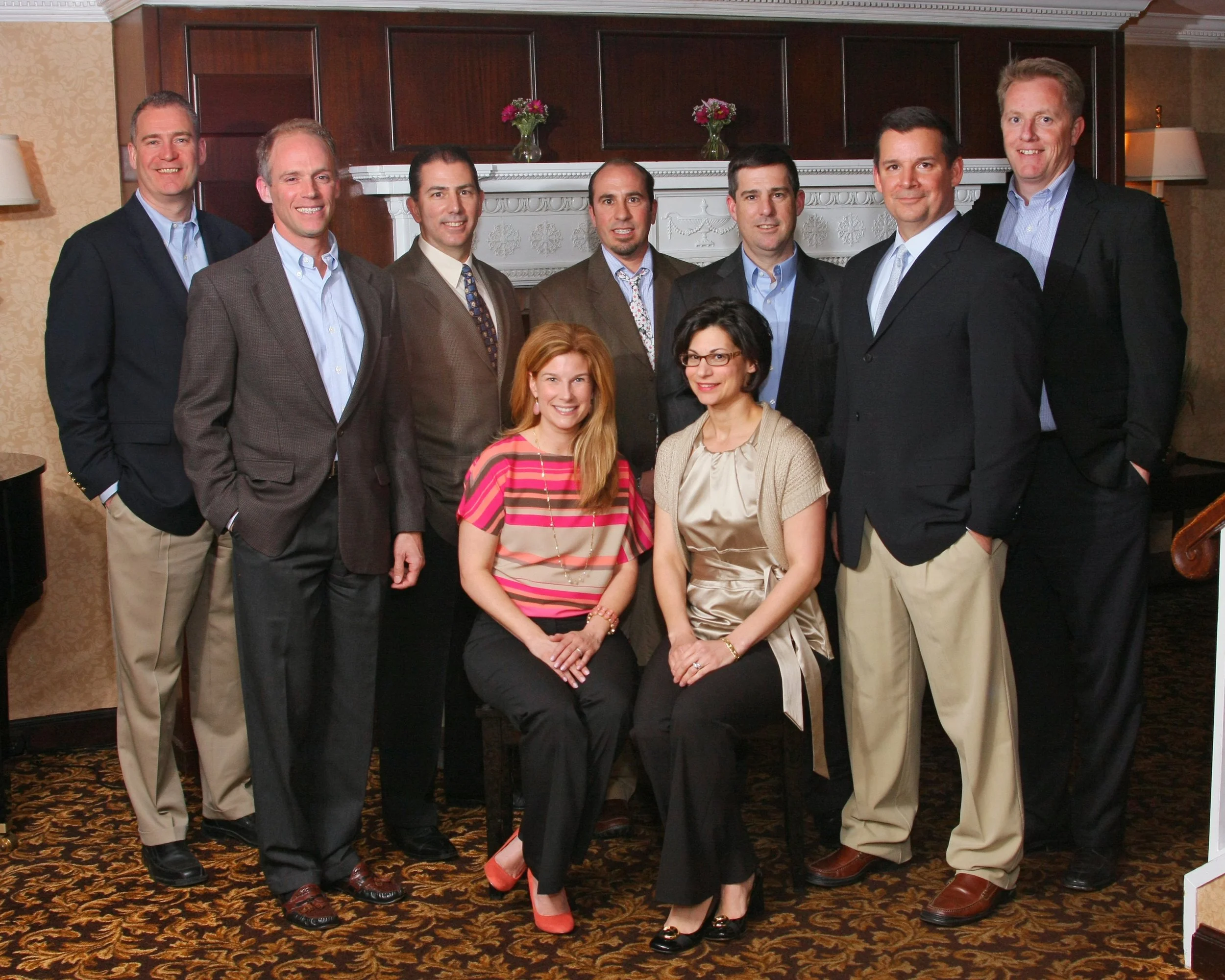 Group of ten professionally dressed people, five men standing in the back row and two women sitting in front, posing for a photo in a formal indoor setting with a fireplace and floral decorations at the Ramsey Golf and Country Club.