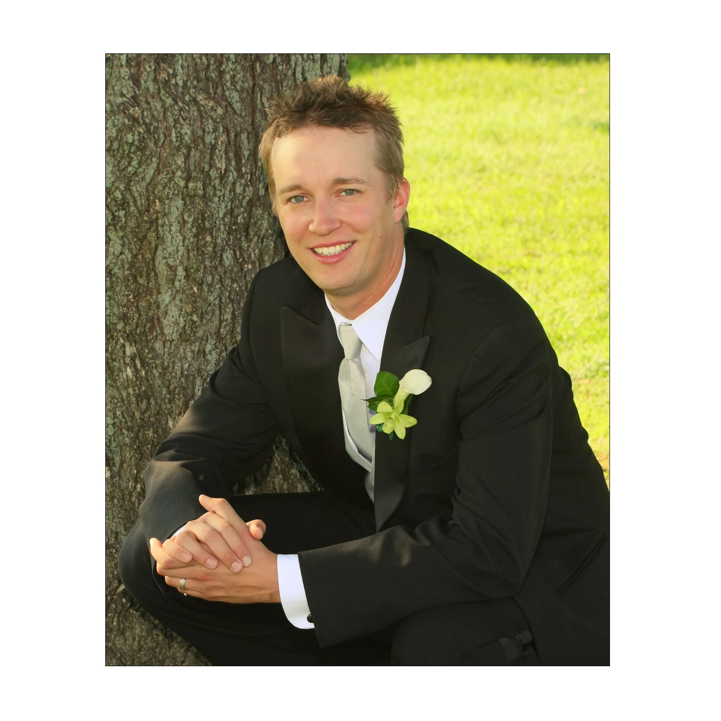 A man in a black suit and white tie sitting outdoors against a tree, smiling, with a boutonniere on his lapel.