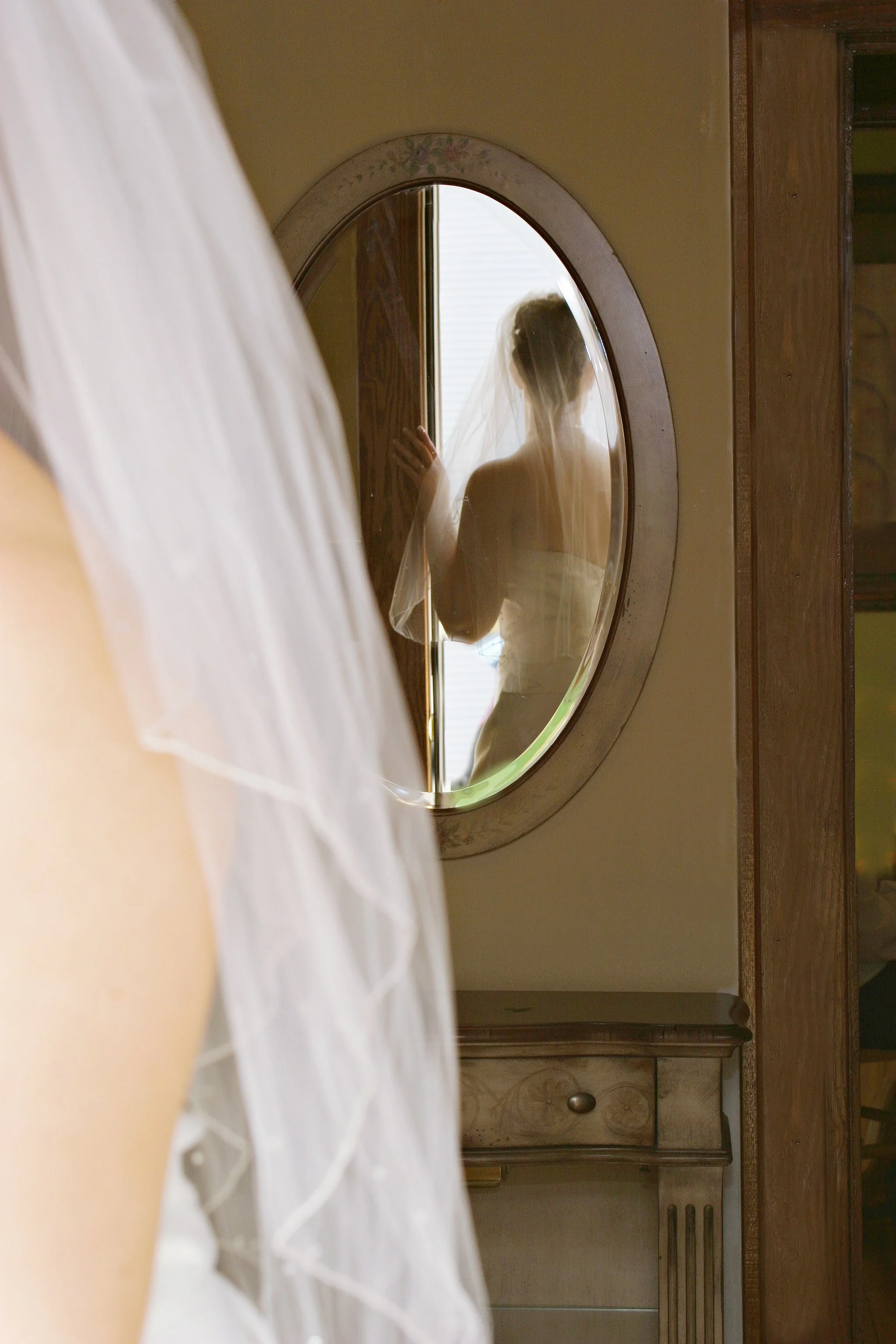A bride standing in front of a mirror, wearing a wedding dress and veil, with her reflection visible in the mirror.