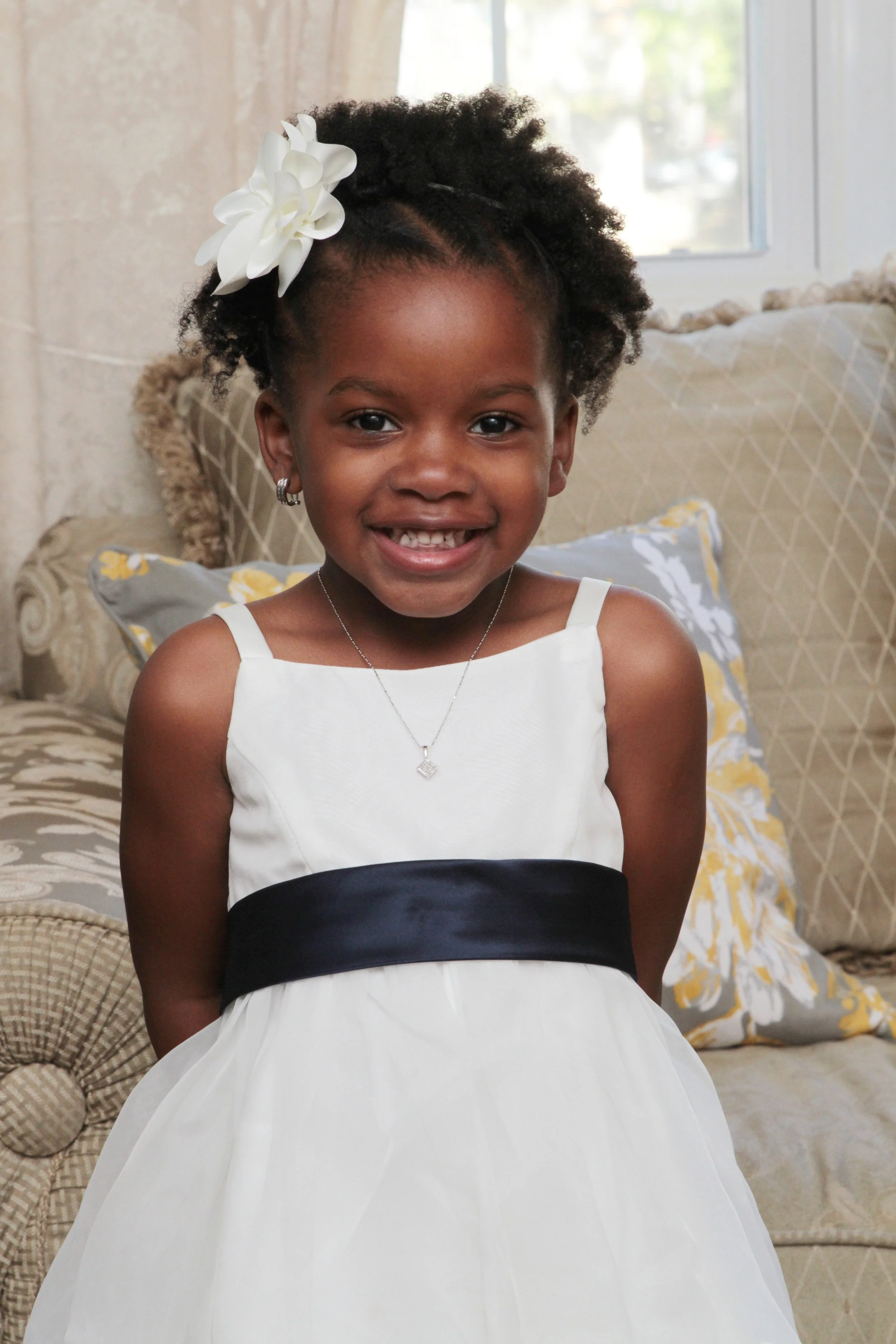 A young girl with a big smile, wearing a white dress with a black sash, silver earrings, a necklace, and a large white flower in her hair, sitting on a sofa in a living room.