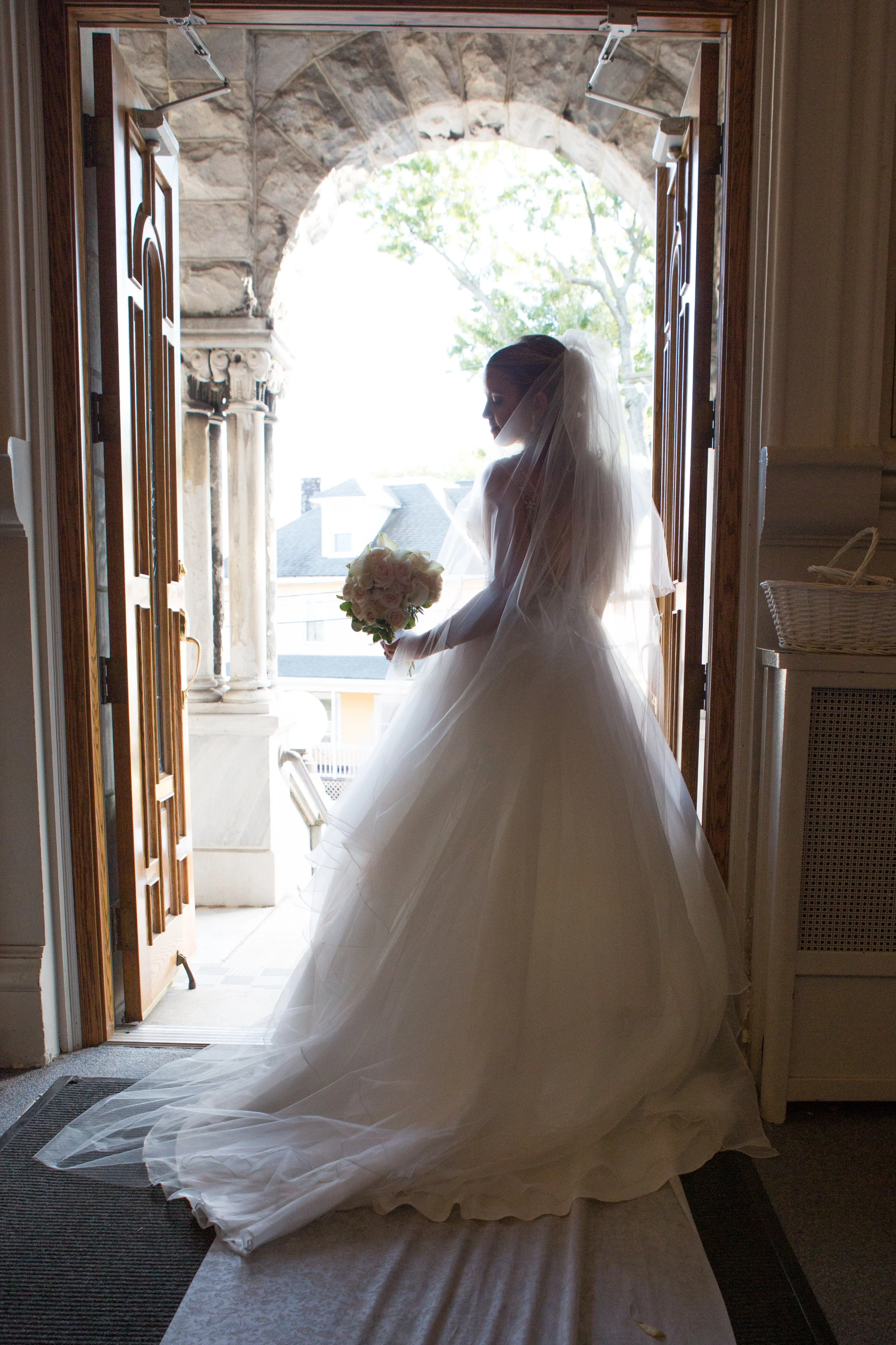 Bride in wedding dress holding a bouquet standing at church entrance, backlit by daylight outside.