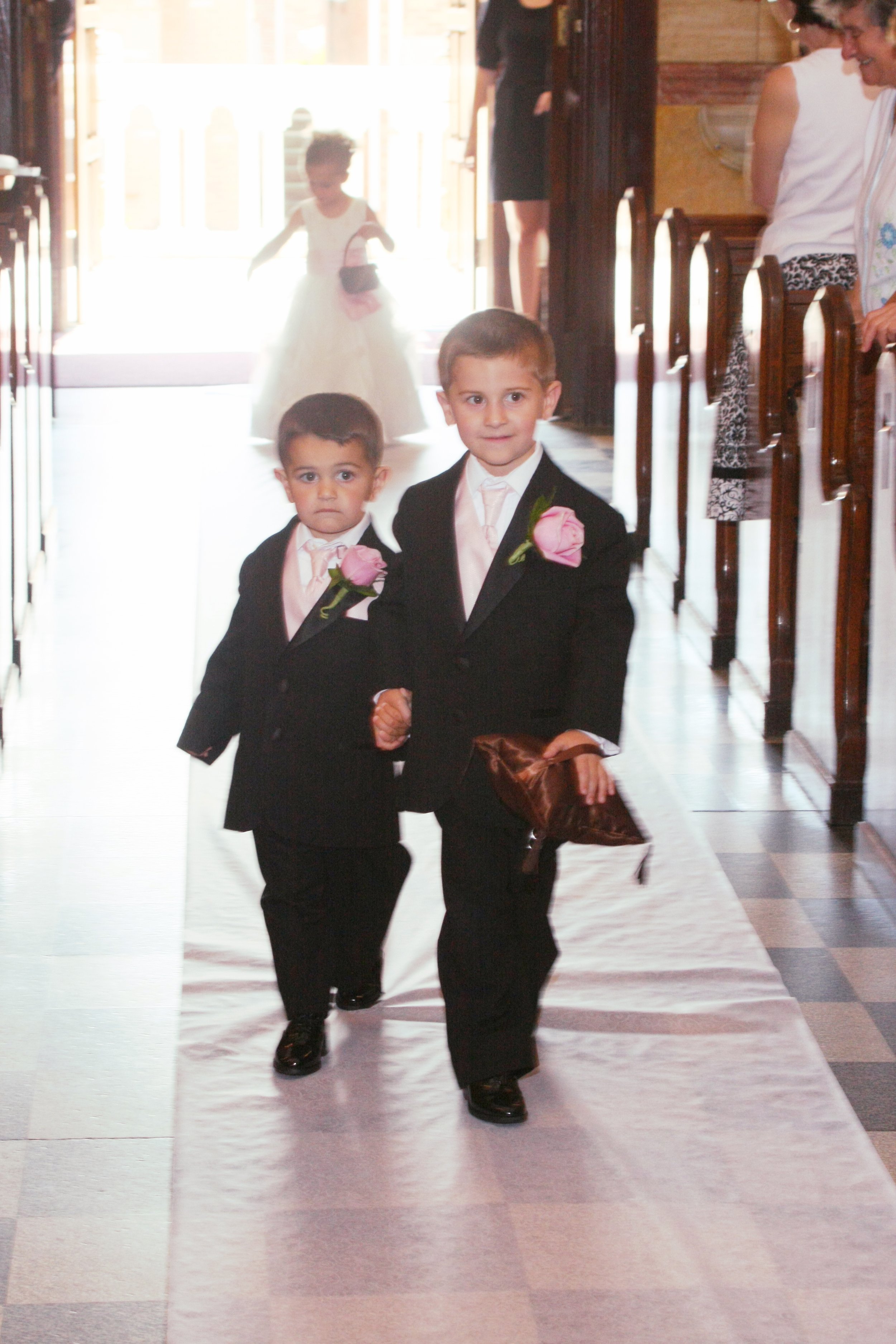 Two young boys dressed in black suits with pink boutonnieres walk down an aisle holding hands at a wedding. A girl in a white dress is visible in the background, and wedding guests are seated along the aisle.