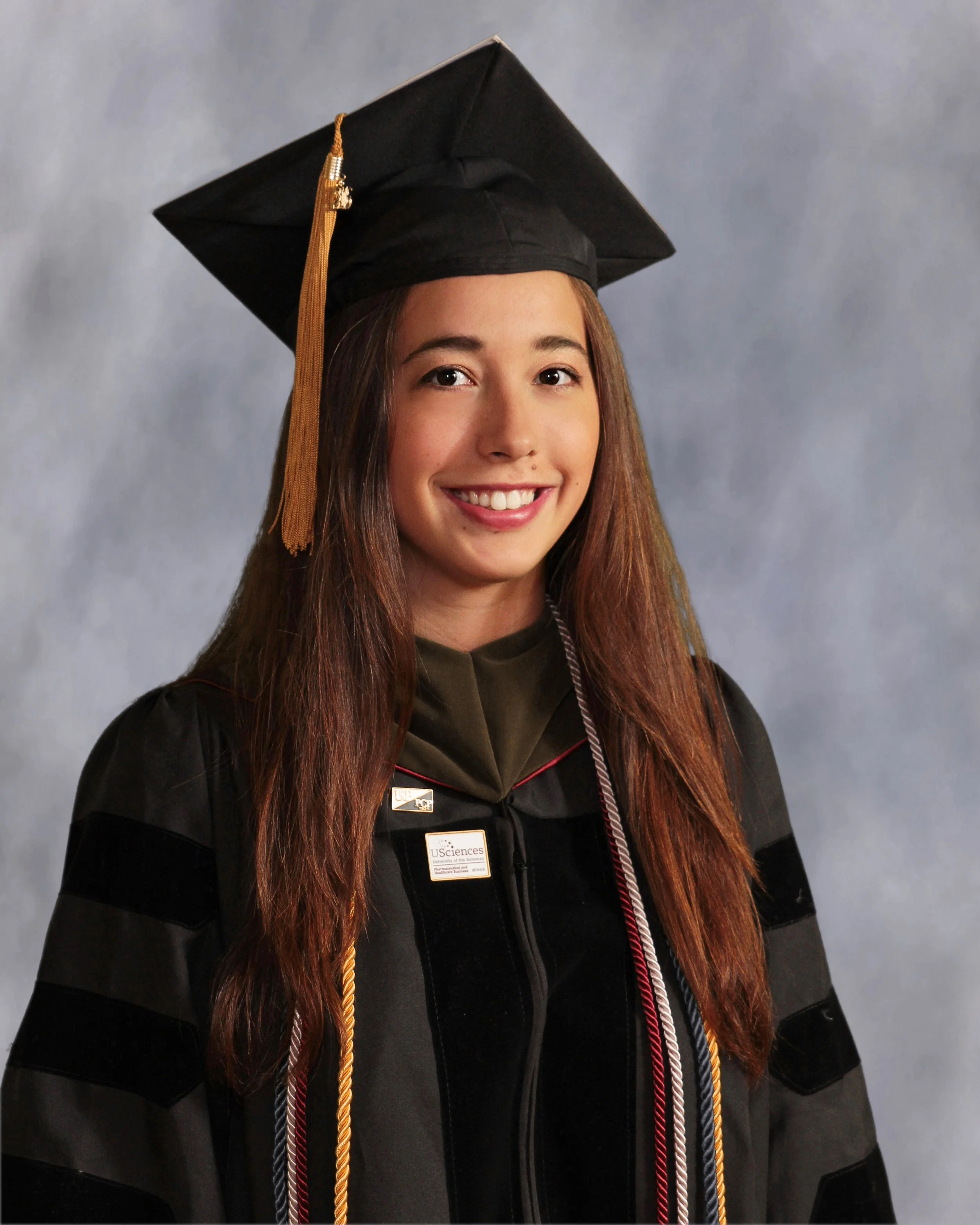 Young woman in graduation cap and gown smiling at the camera, standing against a neutral background for college graduation portrait.