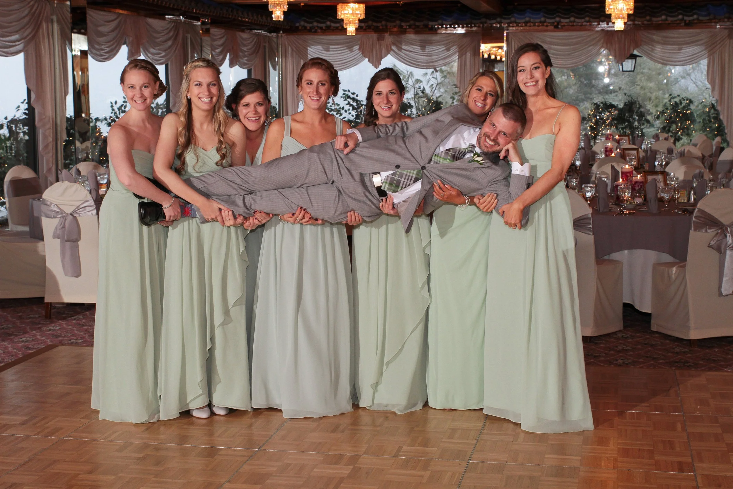 Group of women in matching light green bridesmaid dresses lifting a man in a gray suit and plaid tie on a dance floor at a wedding reception.